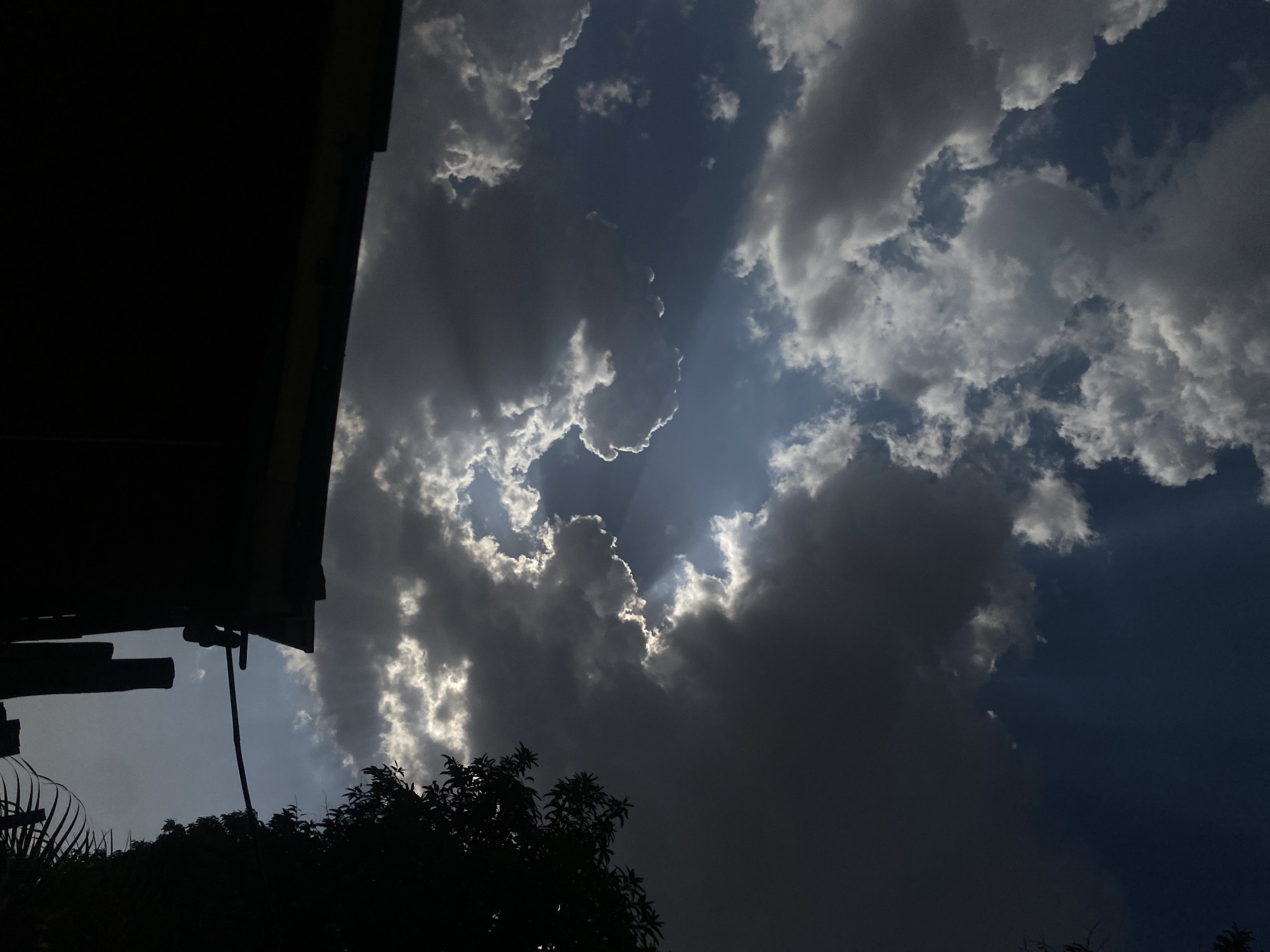 The image, potentially taken in Barden Yana, Nigeria, presents a sky filled with a mix of dark and light clouds, with some rays of sunlight piercing through. The lower portion of the frame includes silhouetted trees and a structure, possibly part of a building.