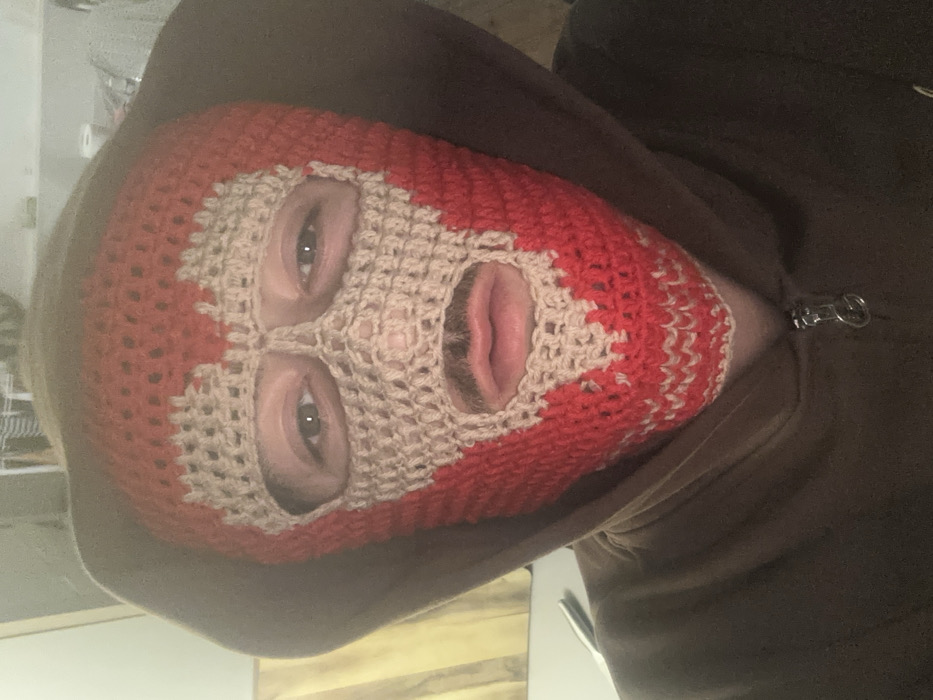 The image depicts a close-up, frontal view of a single individual in an indoor setting, located in the city of Carnin, France. The subject wears a dark brown hooded sweatshirt with the hood pulled up, fully covering the head. A crocheted balaclava covers the face. This balaclava is primarily red, featuring a complex light beige knit pattern that outlines the eye openings, defines a central nose bridge, and frames the mouth opening. Through the balaclava's openings, light-colored eyes are visible, along with a mustache above slightly parted lips. The individual is positioned directly facing the camera. The background is partially visible and out of focus, revealing a light-colored wall or vertical surface on the left. A metallic object, possibly a utensil, rests on an unseen surface to the lower left. Indistinct fabric or material is visible in the upper right background.