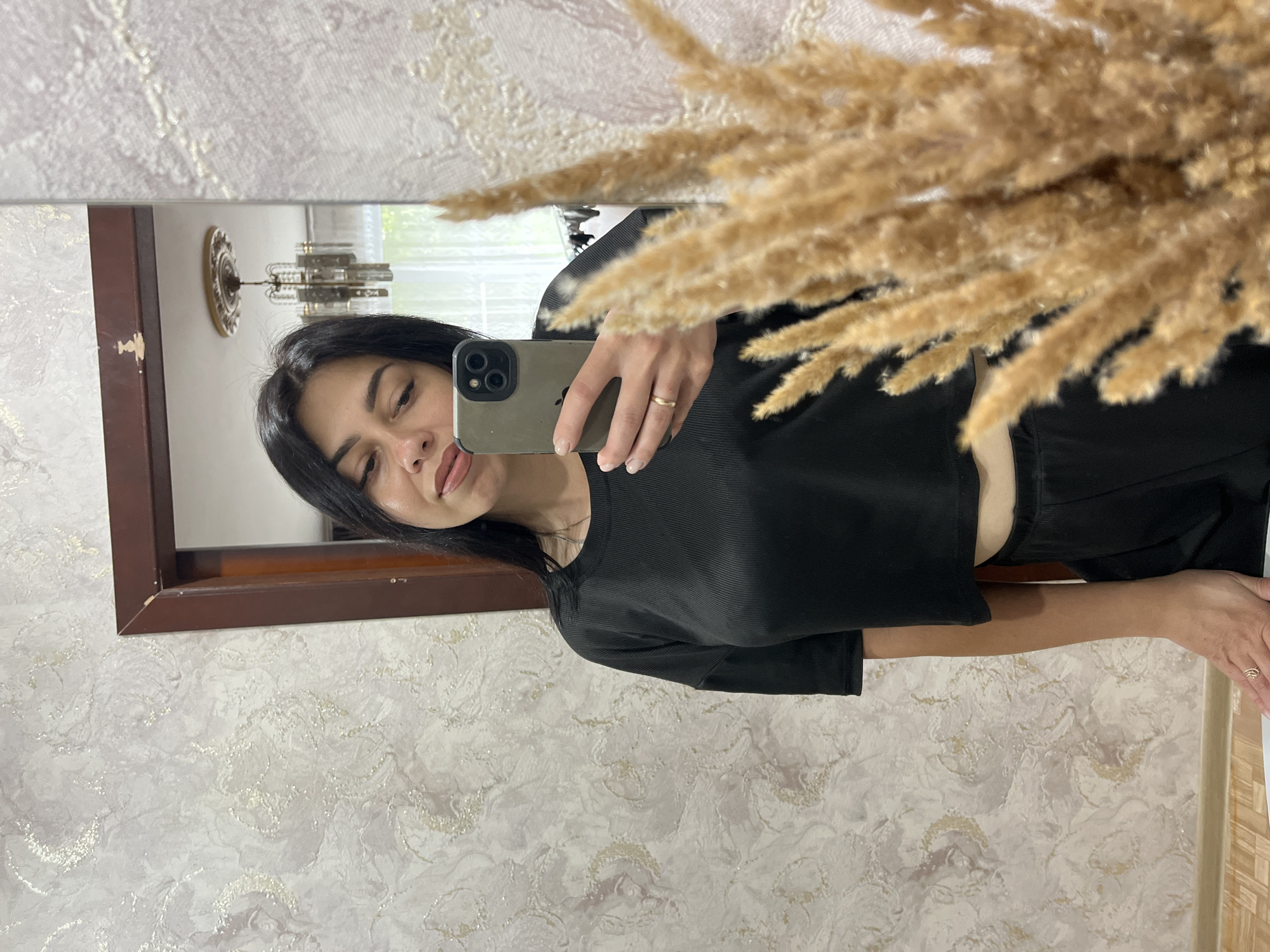 A woman with dark hair takes a selfie in a mirror. She is wearing a black cropped t-shirt and black pants. She has a ring on her left hand. In the mirror's reflection, a chandelier is visible. In the foreground, a decorative dried grass arrangement is visible. The woman is in an indoor setting with patterned wallpaper.