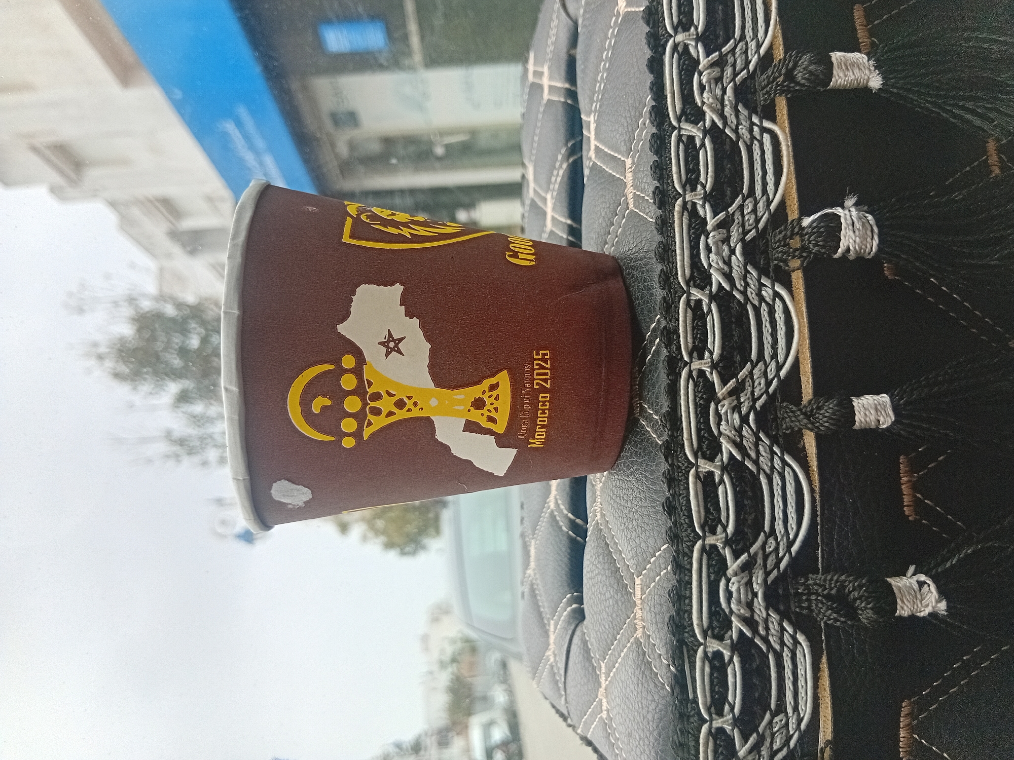 A close-up indoor shot shows a brown paper cup with a graphic of the continent of Africa, the outline of Morocco in white, and a golden trophy, labeled "Africa Cup of Nations Morocco 2025," resting on a black leather car seat cover with white stitching. The cover has a decorative braided trim with tassel-like accents. Outside the car window, a cityscape appears slightly out of focus under an overcast sky, featuring buildings and trees; judging by the cup details, the location is Martil, Morocco.