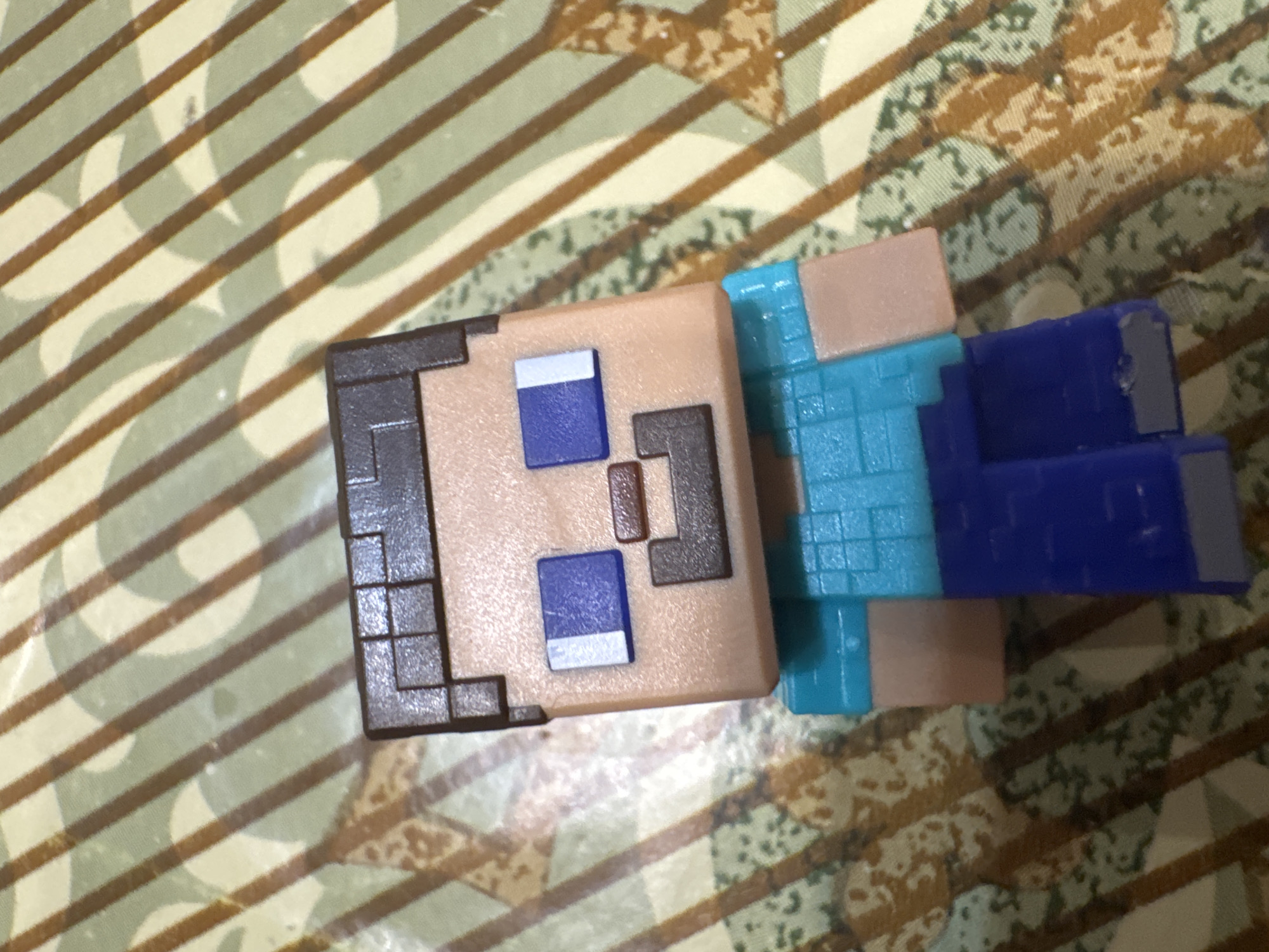The image shows a Minecraft Steve action figure resting on a patterned surface. The figure is composed of blocky elements representing Steve's head, torso, and limbs. The head features pixelated details for hair, eyes, and mouth. The torso and limbs are constructed from blocks of blue and tan plastic. The surface below the figure is decorated with a repeating pattern of beige, white, and brown lines and shapes. Location and country cannot be determined from the image.