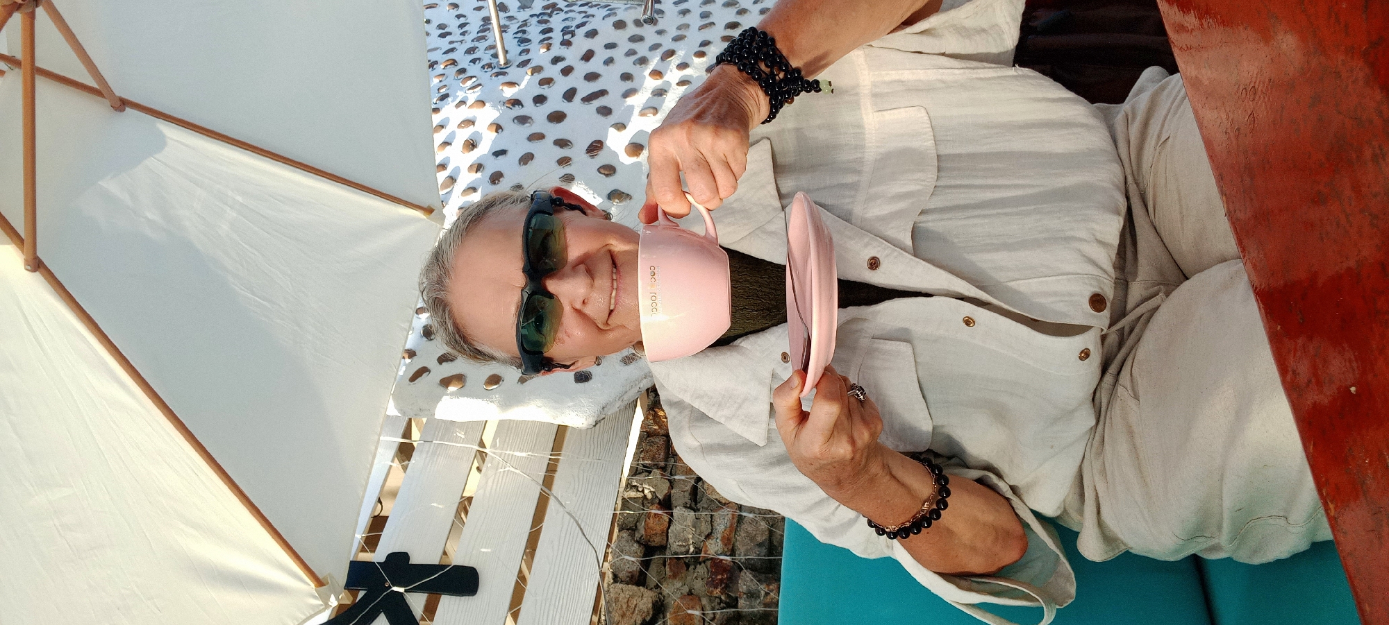 The media file shows a person reclining on a white, slatted surface, possibly a bench or lounge chair, outdoors. A white umbrella provides shade. The person is wearing sunglasses and a light-colored, button-down shirt. They are holding a pink mug in their right hand and a pink saucer in their left hand, appearing to be in the process of drinking or having a beverage. The person is smiling, suggesting a relaxed or content mood. The setting appears to be a sunny outdoor location, likely during the day, with a glimpse of rocky ground and possibly a body of water in the background. Text on the mug reads "cocorococo". The overall impression is one of leisure and enjoyment in a warm climate.