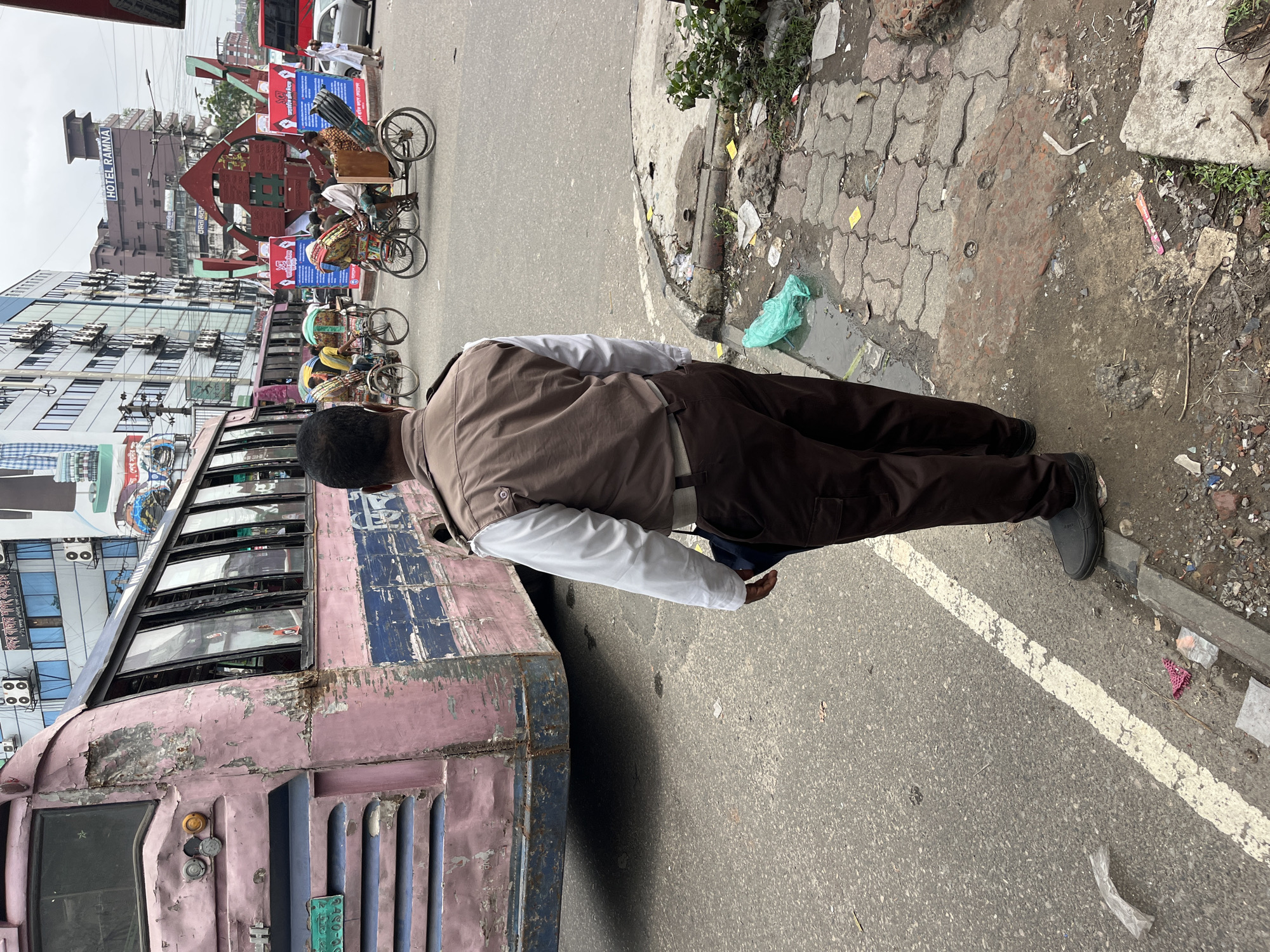 The image captures a bustling street scene in an urban environment, consistent with the provided context of Dhaka, Bangladesh. In the foreground, a person is seen from the chest down, wearing a brown vest over a white long-sleeved shirt, dark pants, and black shoes. They are standing near a large, aged bus that occupies the left side of the frame.
The bus is predominantly pink and blue, but its paint is significantly chipped, faded, and marked with rust, indicating heavy use and age. It features multiple small windows along its side. On a green plate near the front of the bus, the numbers "26080" and "80" are visible. A small sign partially visible near a bus window appears to read "Gazi".
Beyond the bus and the foreground person, the street is active with various forms of transportation and people. Several cycle rickshaws are prominent; one is loaded with long, dark pipes or rods, while others carry passengers. In the mid-ground, a white car is visible, and people are standing or walking nearby.
To the left, a multi-story building with "HOTEL RAMNA" clearly inscribed on its upper section stands tall. Another modern-looking building with numerous windows is also visible further back. A large, abstract, reddish-brown structure or monument is centrally located in the background, partially obscured by blue and red billboards or signs, which display text in Bengali script.
The ground consists of asphalt road and a sidewalk paved with interlocking concrete blocks. The sidewalk and roadside areas show signs of wear and contain litter, including a bright green plastic bag and various debris, suggesting an active public space. The scene is illuminated by bright daylight, suggesting a clear or lightly overcast day. The overall impression is one of typical daily life in a vibrant, busy South Asian city.