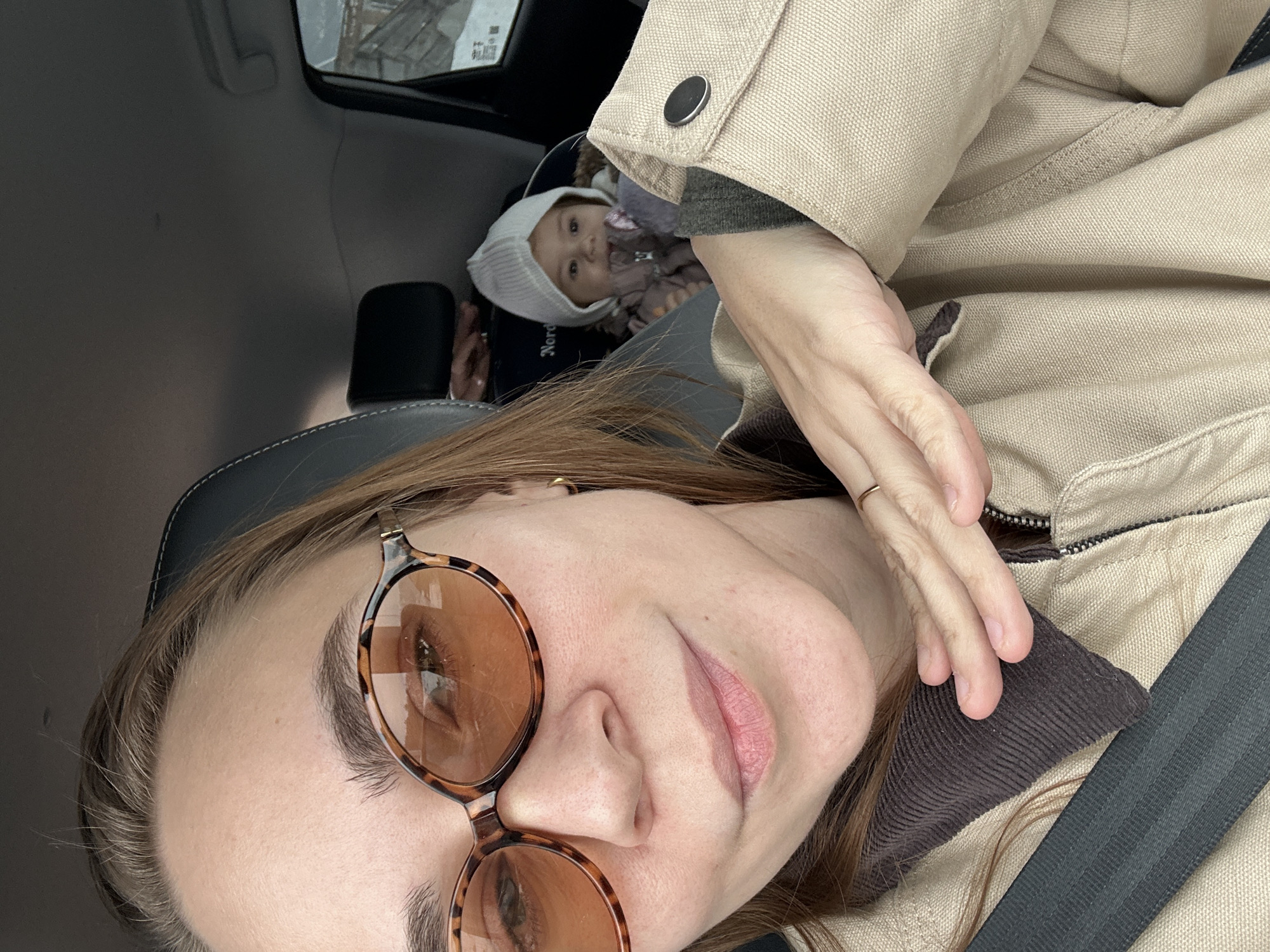 The image shows a selfie of a woman wearing round sunglasses and a beige jacket, positioned in the driver's seat of a car. A baby in a white hat is visible in a car seat in the back. The location is suggested to be Kharnavy, Russia, though visual cues to verify this are not present in the image.