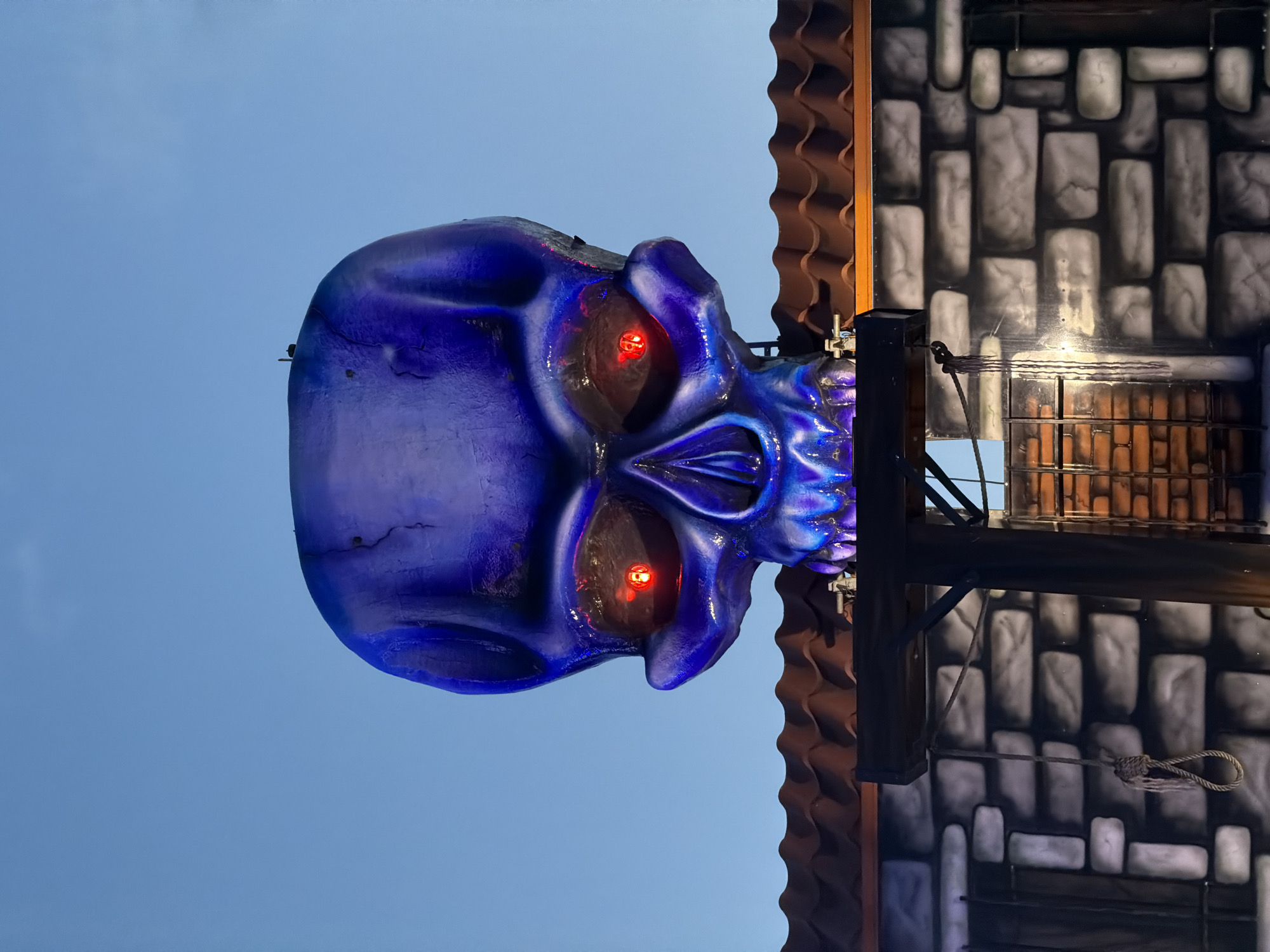 The image shows a large, blue, illuminated skull decoration with glowing red eyes, positioned above a building with a tiled roof and a brick-patterned facade. The skull is made of a textured material, possibly fiberglass or plastic, and has a sinister appearance. Below the skull, a rope with a noose is visible, hanging from a wooden beam. The facade of the building appears to be painted to resemble stone bricks. The scene is set against a clear, twilight sky, with the blue hues of the sky and the skull's illumination creating a dramatic effect. This skull decoration is located at the entrance of the "Horror Alley" attraction at the Sofia Star amusement park in Sofia, Bulgaria.