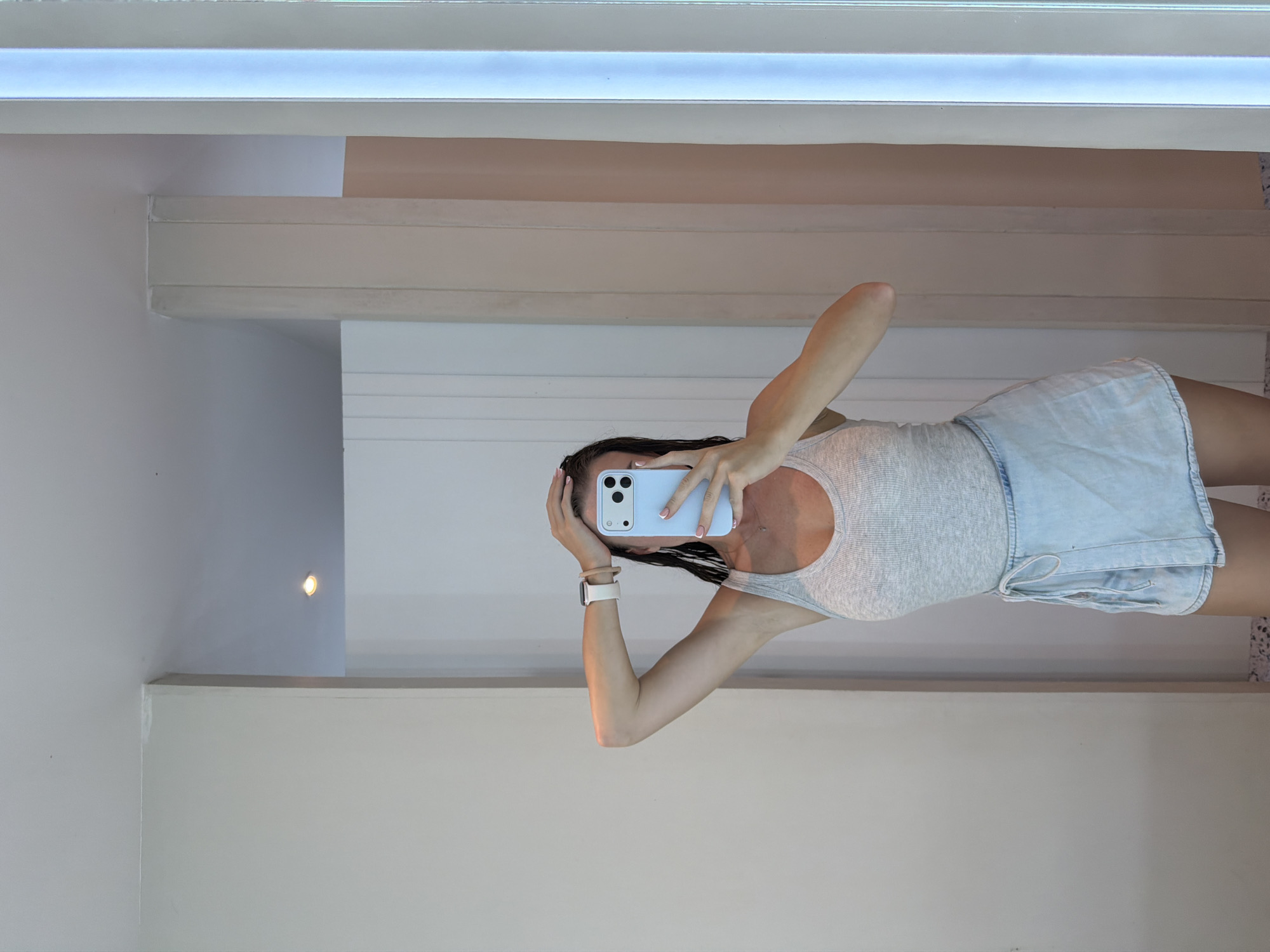 A person is taking a selfie in an interior space, which is presumably located in Ungasan, Indonesia. The person is wearing a light-colored tank top and blue denim shorts. They are holding a light blue smartphone up to their face, obscuring part of their features. Their left hand is holding their hair up behind their phone, and their right hand is covering their face. A white smartwatch is visible on their left wrist.
The background features interior architectural elements, including white walls, a ceiling with recessed lighting, and wooden beams. The lighting appears to be a combination of natural and artificial sources. A small, round light fixture is visible on the wall in the background. The space appears to be a well-lit, modern interior.