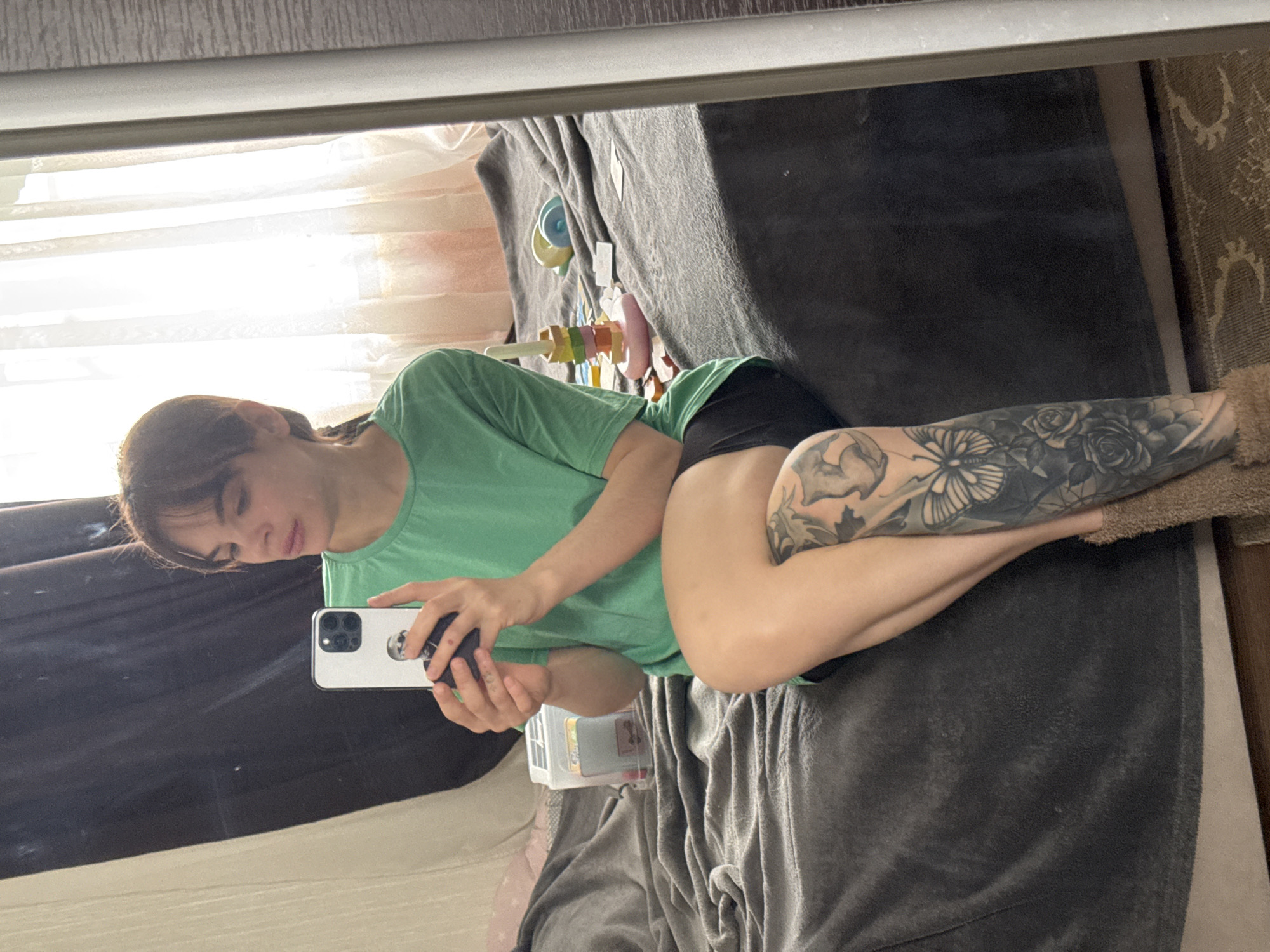 A mirror selfie captured indoors in Rostov-on-Don, Russia, shows a young person lying on their side on a dark grey, textured blanket or duvet. The individual, appearing female, has dark hair with bangs and is wearing a light green t-shirt and black shorts. They are holding a white smartphone, likely an iPhone, in both hands, looking at the screen as they take the photograph.
A prominent and detailed black and grey tattoo featuring floral and butterfly motifs covers much of their visible right leg (the leg closest to the viewer). The background reflects a brightly lit window with white sheer curtains, indicating daytime. Various colorful objects, including what appears to be a wooden toy with colored blocks and other decorative items, are visible on a surface behind the person. A patterned rug is also partially visible on the right side of the frame, contributing to the cozy, indoor setting. The person's expression is neutral, focused on the phone. No discernible text is visible on any objects.