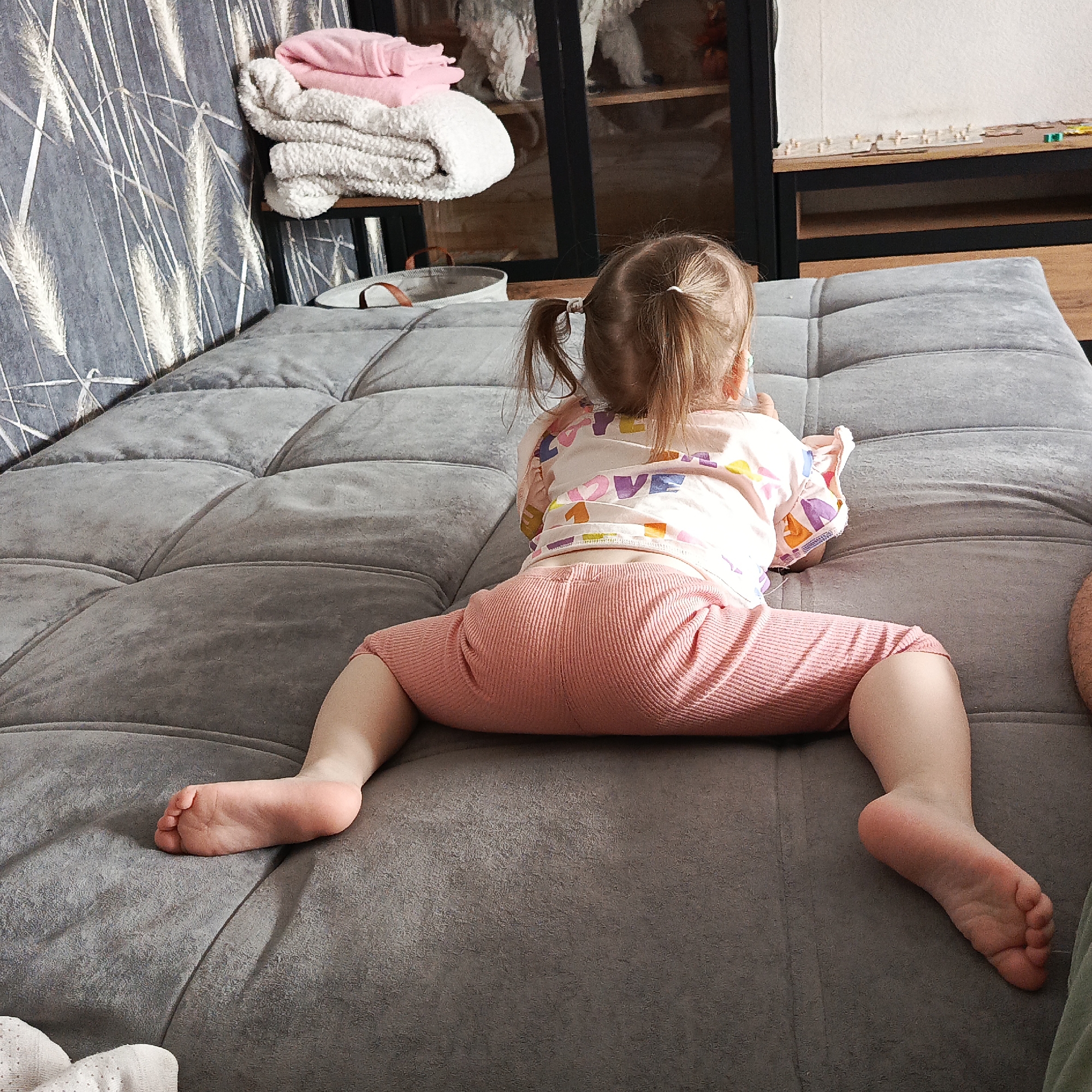 A young child is seen from the back, seated on a gray, cushioned sofa. The child wears a light pink shirt with colorful letter and shape designs and darker pink ribbed pants. The child's legs are spread wide, feet pointing outwards. The child has light brown hair tied into two small pigtails. A partially visible adult leg in green fabric appears on the right side of the frame. The location is identified as Марковский, Russia, based on metadata. In the background, a sofa with a patterned cushion is visible to the left, and a cabinet with a display of decorative objects stands behind the sofa. A table with small items on it can also be seen.