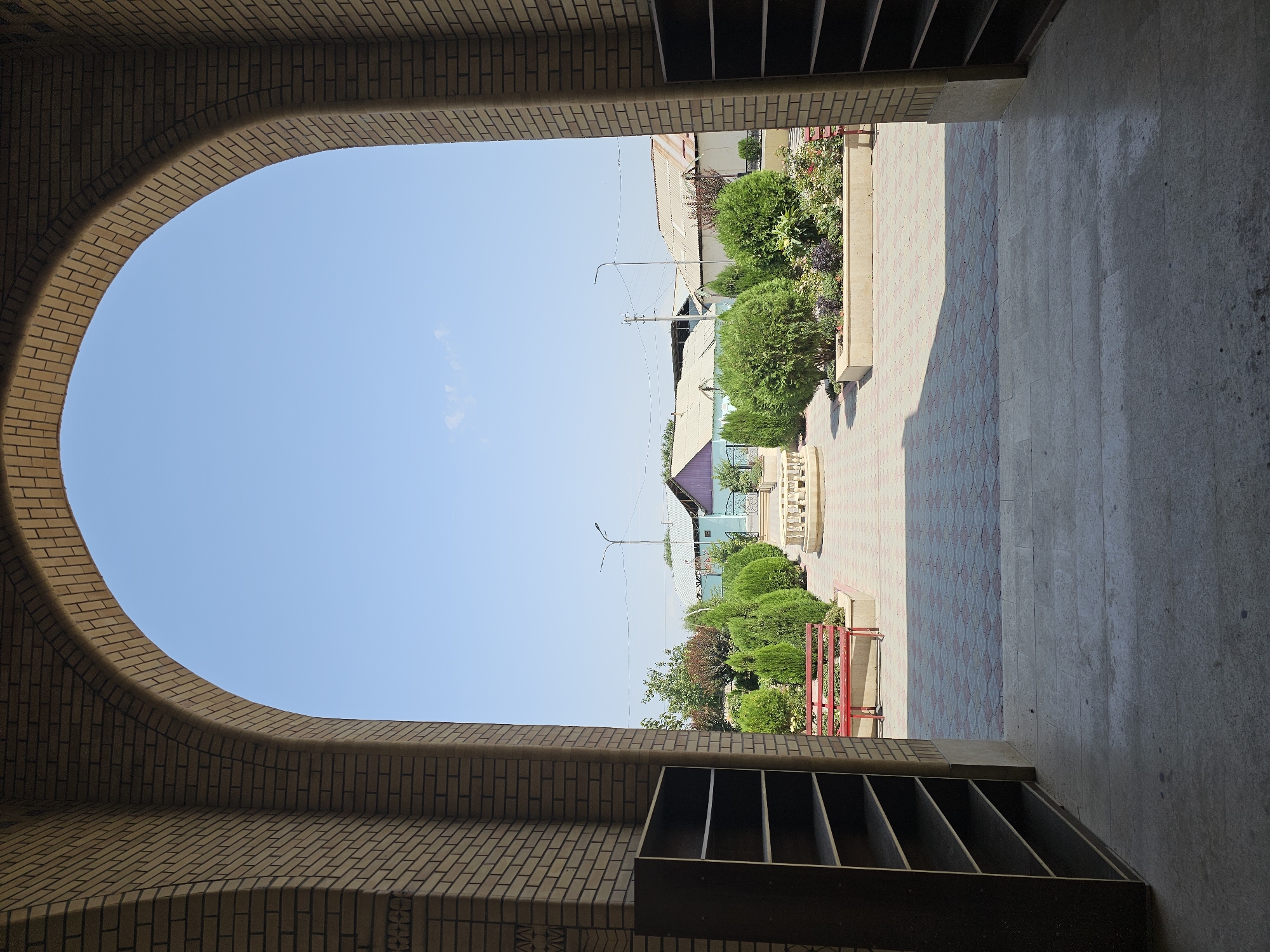The image shows a view through a large brick archway, framing a sunny courtyard. On the left and right sides of the archway are tall, dark brown shelving units. The courtyard has a patterned brick pathway leading to a small fountain in the center. There are several evergreen bushes and some flowering plants arranged in a raised planter. A red bench sits to the left of the pathway. In the background, there are several low-rise buildings with pitched roofs, and a street lamp. The sky is clear and blue. The scene appears to be within the city of Özgön, Kyrgyzstan, possibly within the grounds of a cultural or religious site, given the architectural elements and the serene courtyard. There are no people visible in the image.
