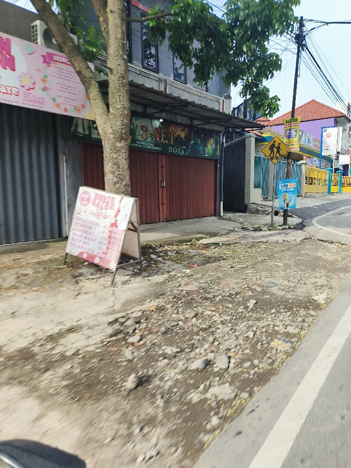 The image shows a street corner in Soreang, Indonesia, located at latitude -7.018 and longitude 107.544. The scene features a paved sidewalk with a sign for a salon advertising smoothing, cutting, coloring, and hair washing services. In the background, a closed storefront, decorated with graffiti and a sign reading "3-NET - 1 MILLION YOUTHS WORLDWIDE - DOTA 2," is situated on the corner. A telephone pole with a yellow "children crossing" sign stands in front of the storefront. The pavement is uneven and contains many loose rocks and debris. The street appears to be empty of traffic and pedestrians. The image is taken from the perspective of a vehicle driving down the street.