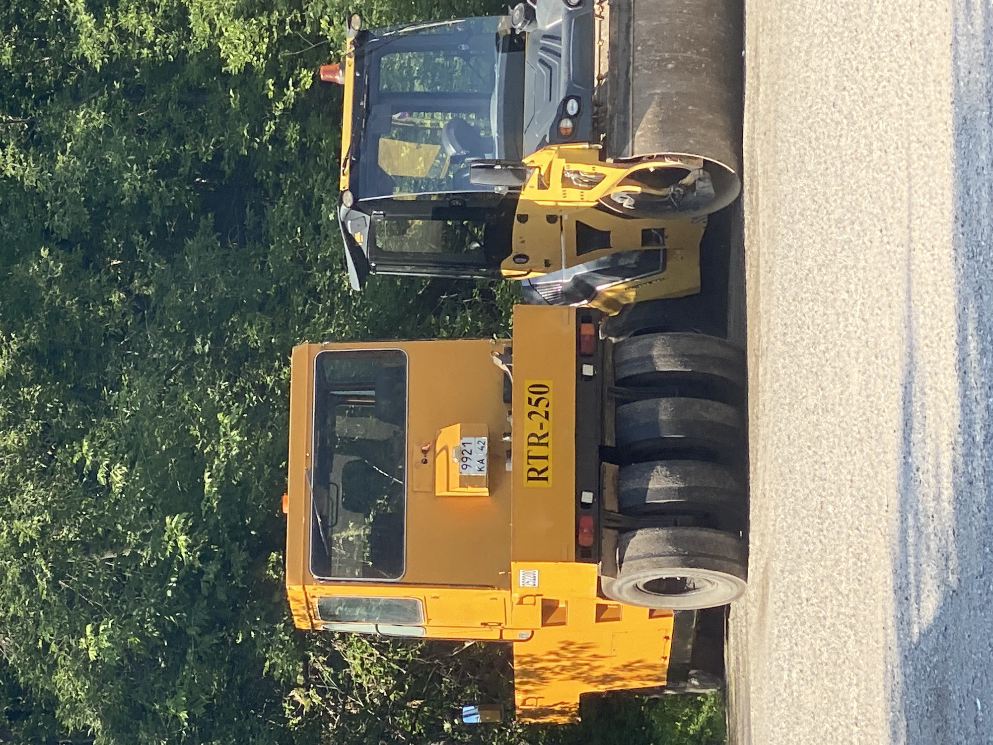 Two industrial vehicles, a yellow tow tractor and a yellow and black road roller, are parked on a gravel surface in front of a dense green tree line. The tow tractor is on the left and has a single cab with a yellow exterior and windows. It has multiple large black tires on the rear. A license plate with the numbers "9921 KA 42" is visible on the back of the tow tractor, along with the text "RTR-250". The road roller is on the right, with a prominent steel drum at the front and a cab with windows and a black roof. An orange flashing light is visible on top of the road roller's cab. The environment suggests an outdoor industrial or construction setting. The specific location in Novokuznetsk, Russia, cannot be determined from the visual information alone.