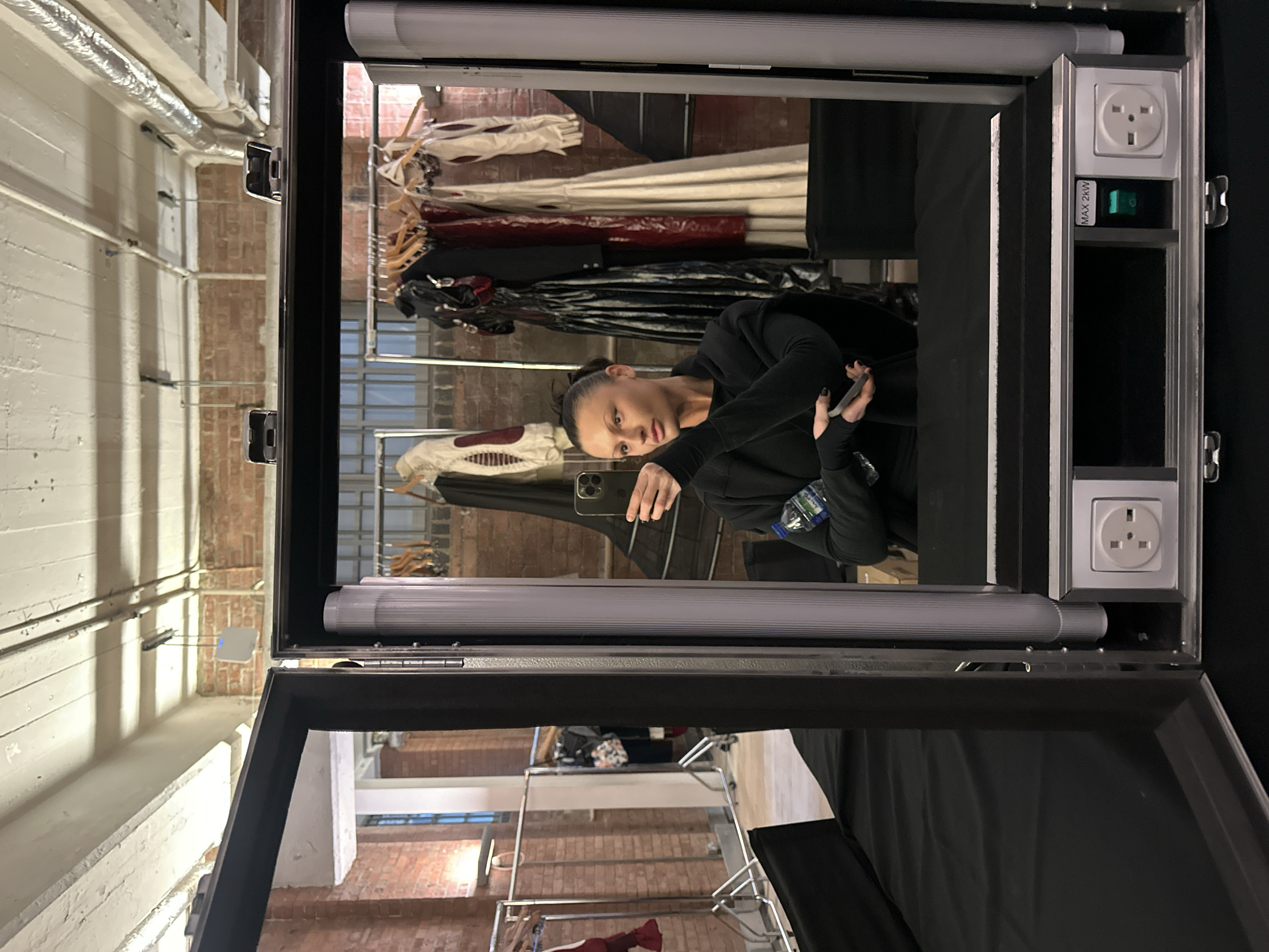 The image depicts a woman's reflection in a portable, illuminated dressing room mirror within an industrial-style interior, located in the city of London, United Kingdom.
The woman, visible from the chest up, has dark hair pulled back and wears a dark, long-sleeved top. She holds a black smartphone with a triple-lens camera array in her left hand, positioned to capture the reflection. Her right hand grasps a clear plastic water bottle and a small dark electronic device. Her fingernails are painted dark.
The portable dressing mirror unit features vertical tubular lights on either side of the mirror. Below the mirror, two white Type G electrical sockets, characteristic of the United Kingdom, are integrated alongside a green illuminated power switch labeled "MAX 2KW." The immediate surface in front of the mirror is covered in black fabric.
The background, visible as a reflection in the mirror, reveals a spacious room with exposed reddish-brown brick walls. The ceiling is characterized by white-painted wooden beams, planks, exposed white pipes, and silver ducting. Large, multi-pane windows are visible further back. Several metal clothing racks are present, holding a variety of garments on wooden hangers, including a light-colored bomber jacket with red elbow patches, long dark dresses, a deep red garment, and a futuristic light-colored outfit with exaggerated shoulders and integrated gloves. Additional silver metal stands and black fabric-covered surfaces are also visible in the reflected environment.