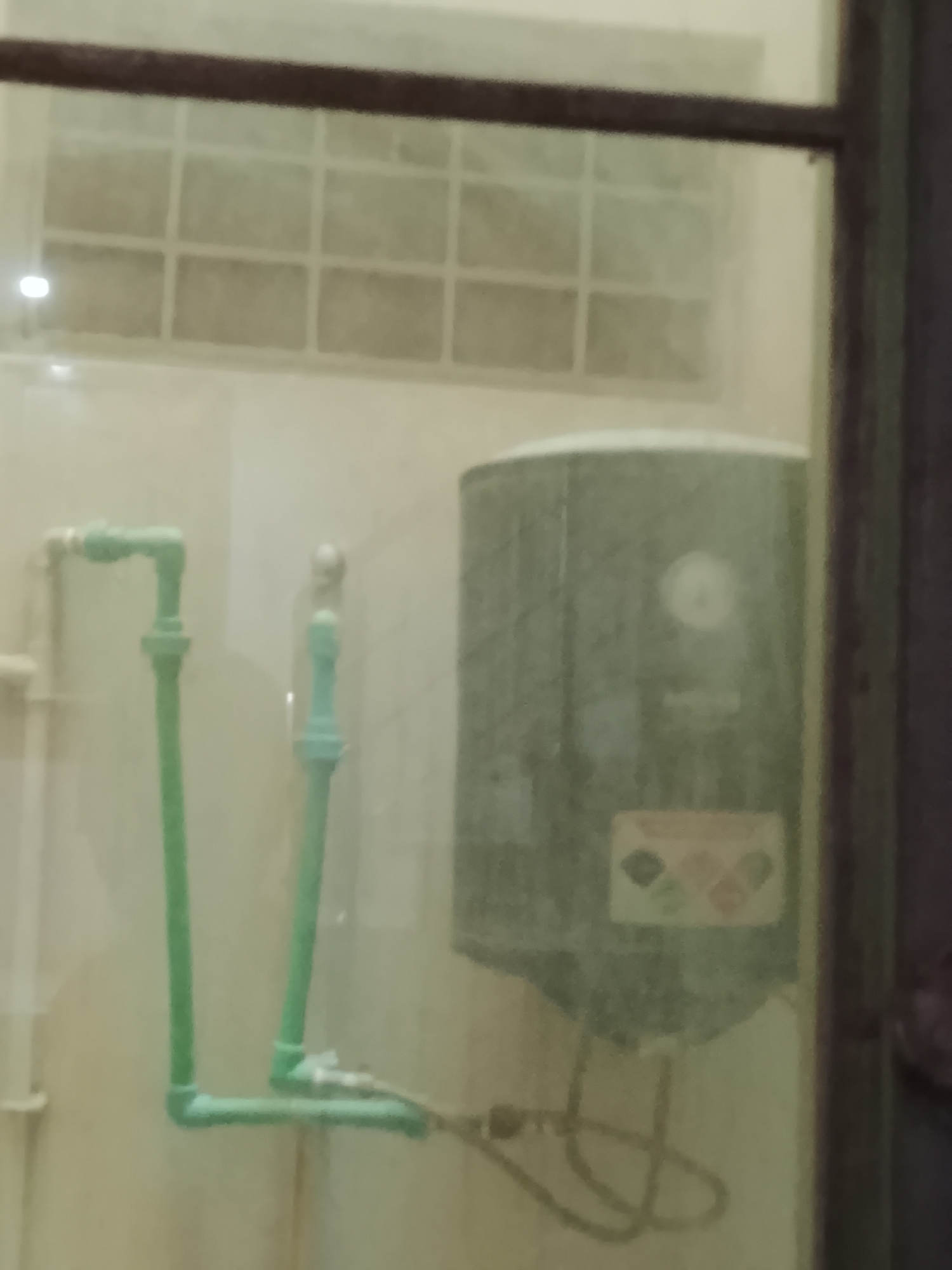 The image appears to be taken inside a building in an unknown city. The scene includes plumbing pipes, a water heater, and a window with a grid pattern. The water heater has a round gauge and a safety warning label. The pipes are green and appear to be made of PVC. The environment is mostly white or off-white. The overall image is somewhat blurry, possibly taken through a window or reflective surface. No people are visible.
