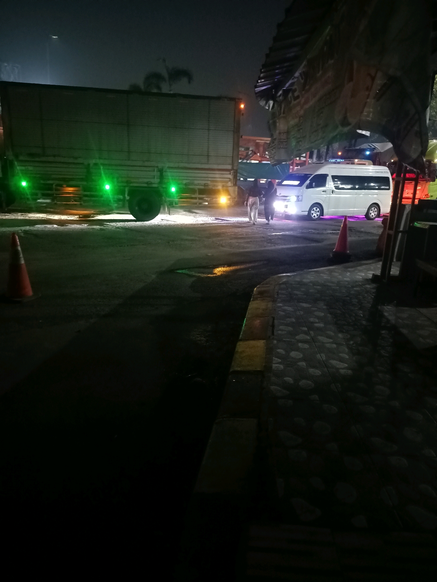 At night, in Karawang, Indonesia, a large truck with green lights is parked on a paved surface, possibly a lot or a road. To the right of the truck, a white van with its headlights on is also parked. Two people are standing near the van. In the foreground, an orange traffic cone is placed on the ground. The scene is dimly lit, with the ambient light coming from the vehicles and some streetlights in the background. The ground appears to be wet or reflective, suggesting recent rain or moisture. The overall atmosphere is urban and industrial.