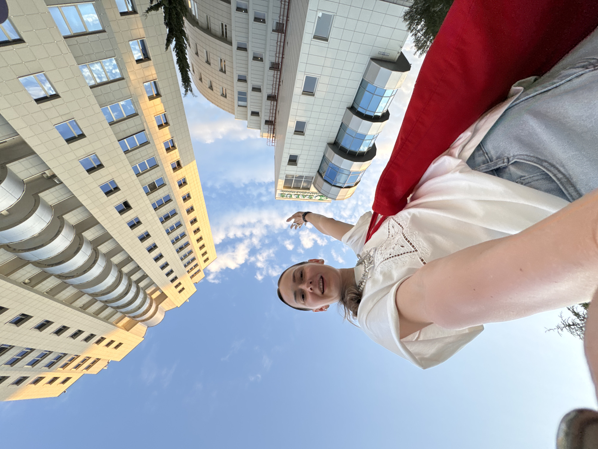 A low-angle, wide shot captures a young woman in a white t-shirt, light blue jeans, and a bright red scarf draped over her shoulder. She stands outside, looking up and towards the camera, with her right arm extended as if waving. Behind her, two modern, multi-story buildings with pale facades and numerous windows dominate the sky. The building on the left features curved balconies, while the one on the right has a more angular design and a prominent glass-fronted section with the text "SALUS" visible on its upper portion. The sky is a clear blue with a few wispy clouds. The sunlight illuminates the buildings with a warm, golden hue, suggesting it is either early morning or late afternoon. The scene appears to be set in a contemporary urban environment, likely a business or residential district. The architecture suggests a location in Sochi, Russia, known for its modern development.