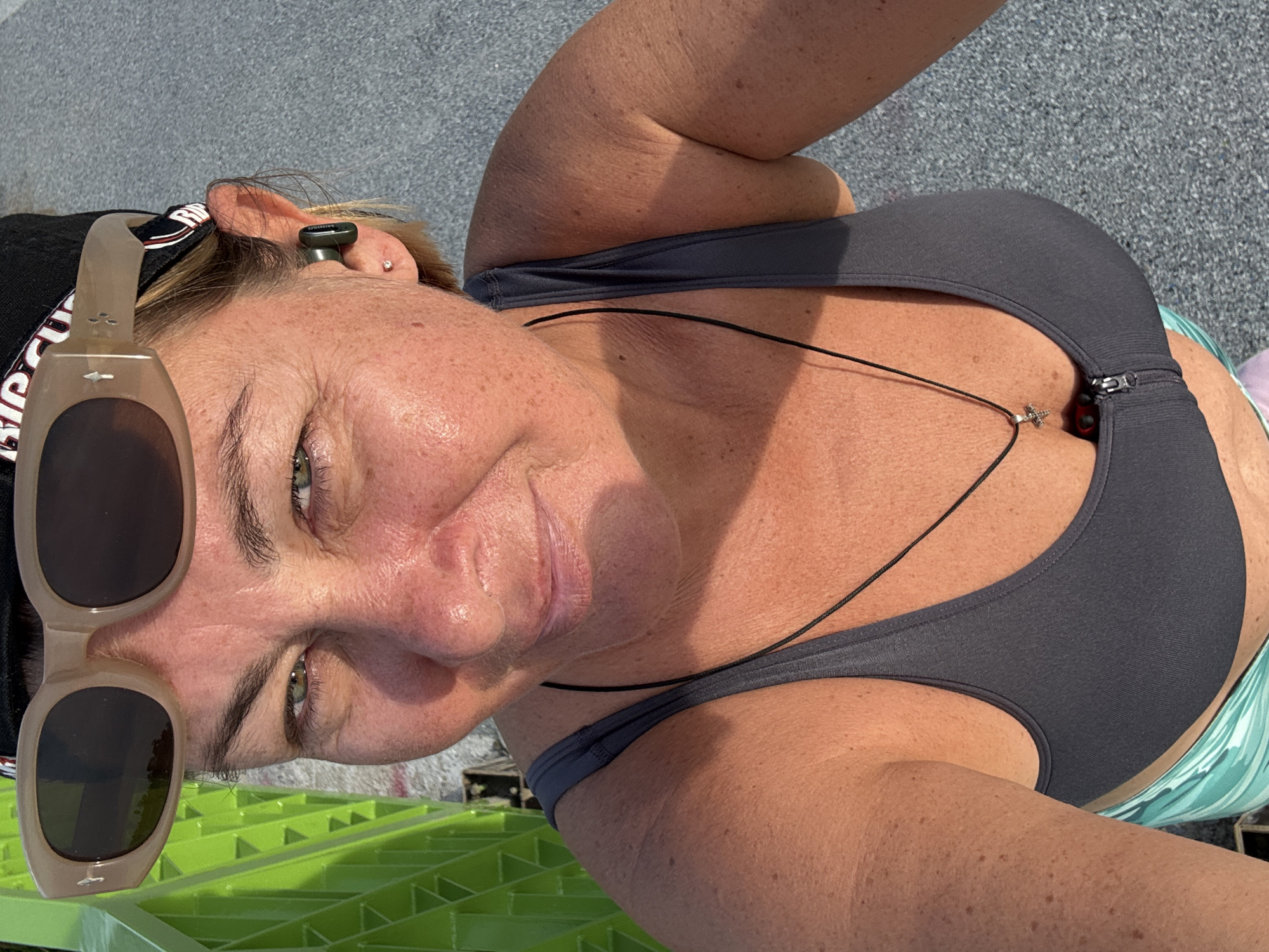 A tanned woman is facing the camera, wearing sunglasses and a black sports bra with a black necklace. The background shows a gray surface, and green plastic. The location and date are unknown.