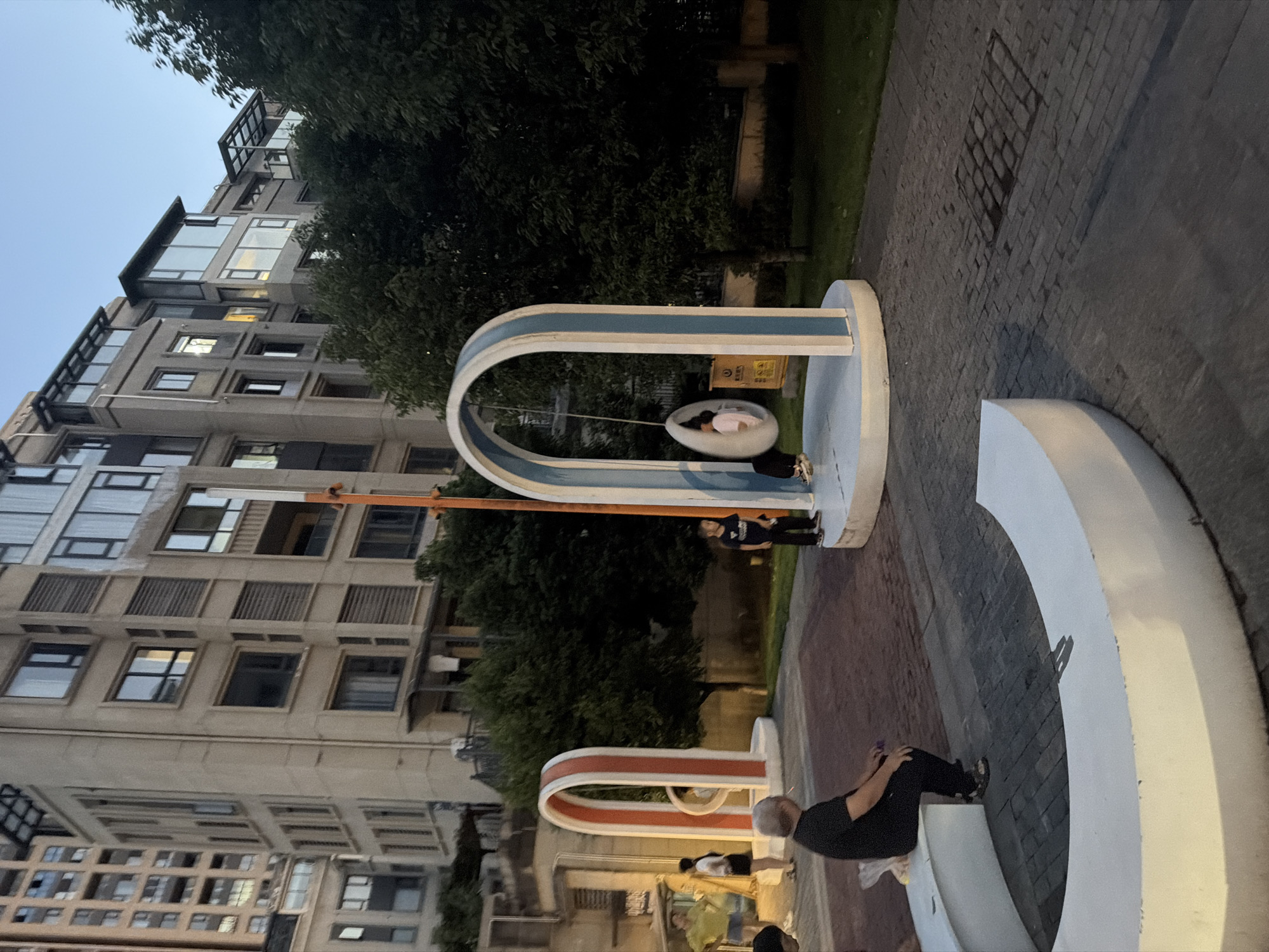 The image depicts an outdoor public space in Jiangjin, China, during dusk. In the foreground, a man in a black t-shirt and pants is seated on a curved white bench, facing away from the camera and towards a plaza. Behind him, a child in a dark t-shirt and pants stands near a modern playground structure featuring an arched frame with blue accents, inside which a person is seated in an egg-shaped swing. To the left, another arched structure with red and white stripes stands on a circular platform. In the background, a multi-story residential building with numerous windows and balconies rises behind a line of trees. The ground is paved with brickwork and stone tiles. Several other individuals are visible in the middle ground, including a woman in a yellow shirt and shorts, and a woman in a white shirt and black shorts. The overall lighting suggests the transition from daylight to evening.