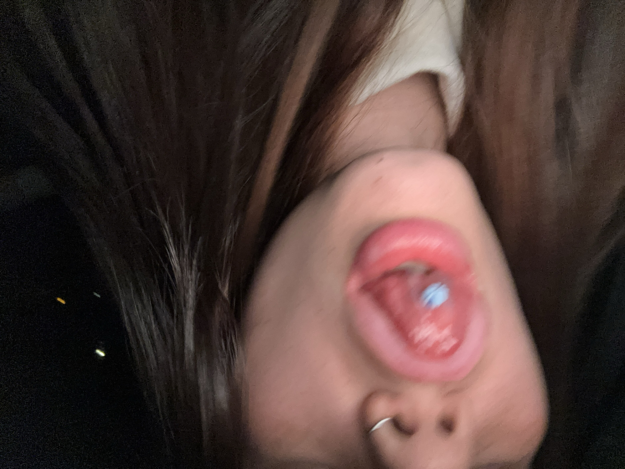 A close-up, upside-down selfie shows a person with brown hair, a silver nose ring, and a blue pill on their tongue. The background is dark. The location is indeterminable from the image.