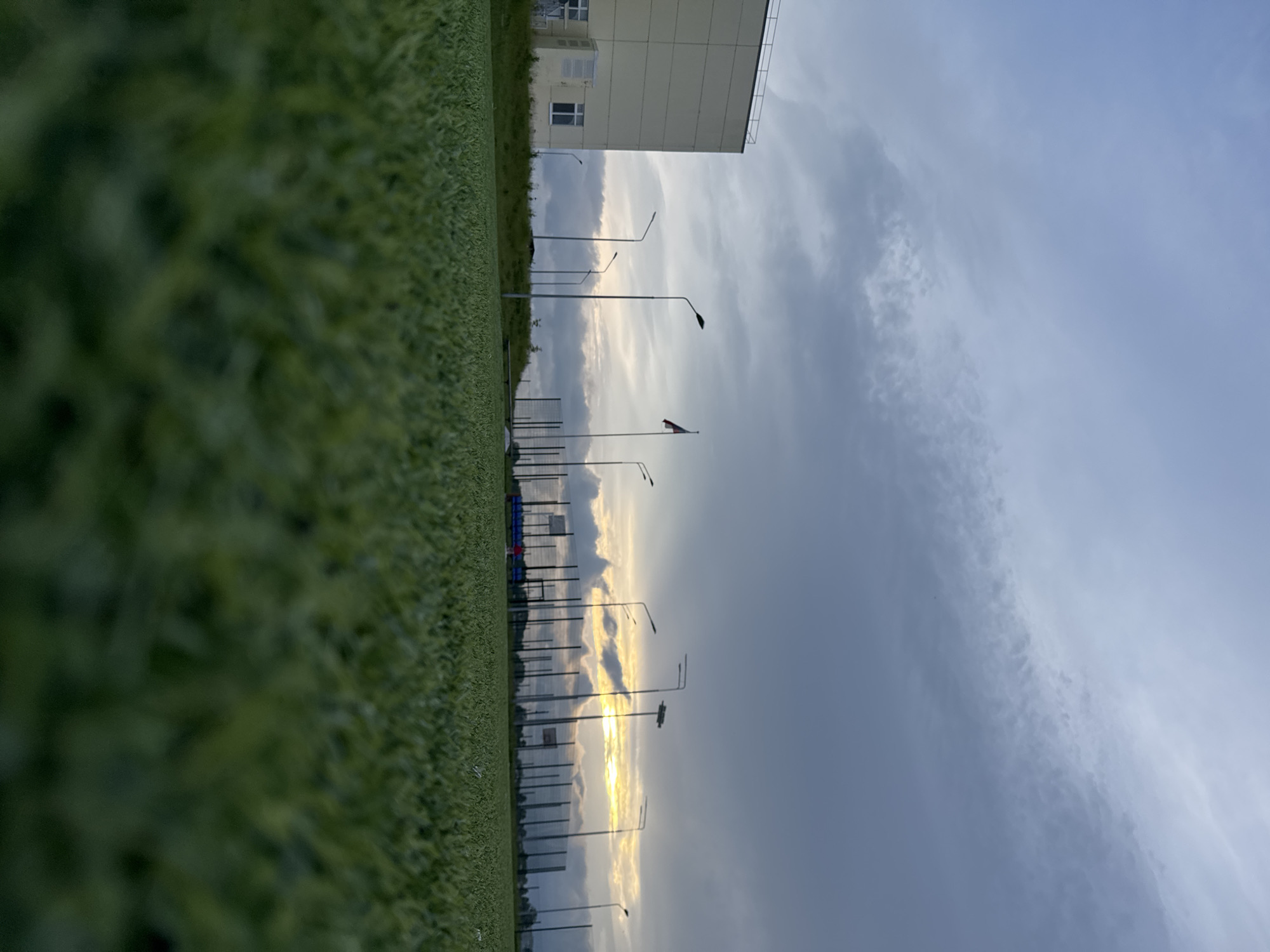 The image captures an outdoor scene in Ферма, Russia, featuring a low-angle perspective of an artificial green turf field that occupies the immediate foreground, gradually sharpening towards the midground. On the left side of the midground, a light-colored, multi-story building with multiple windows and external air conditioning units is visible. Extending across the midground from left to right is a series of tall, dark streetlights or floodlight poles. A tall flagpole displaying a red and white flag, indicative of the Russian national flag, stands between the building and the right side of the frame. Further to the right, a fenced outdoor sports court with a visible basketball backboard is present. Sparse green vegetation grows on a slight elevation behind the infrastructure. The sky in the background is largely overcast with grey and white clouds. A prominent yellow and orange glow, partially obscured by clouds, is visible on the horizon, consistent with either sunrise or sunset. No individuals are depicted in the scene.