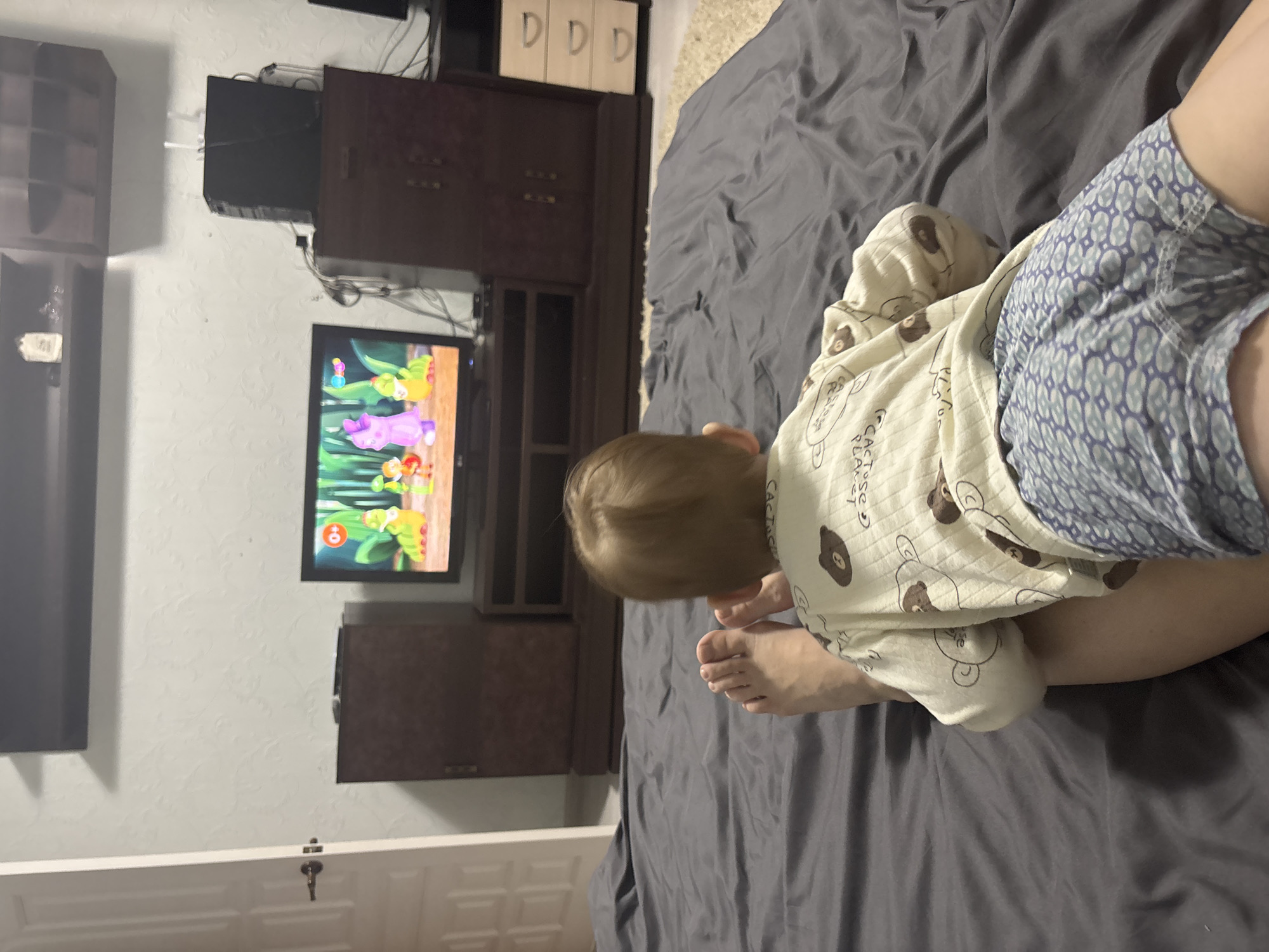A child wearing a beige and brown patterned pajama top and blue and white patterned shorts is lying on a gray bed in a room with a television. The television displays a cartoon. A dark wooden entertainment center is behind the television. Shelving and cabinets are visible. The room's wall is white. A white door is visible in the lower left. The location is Ferma, Russia.