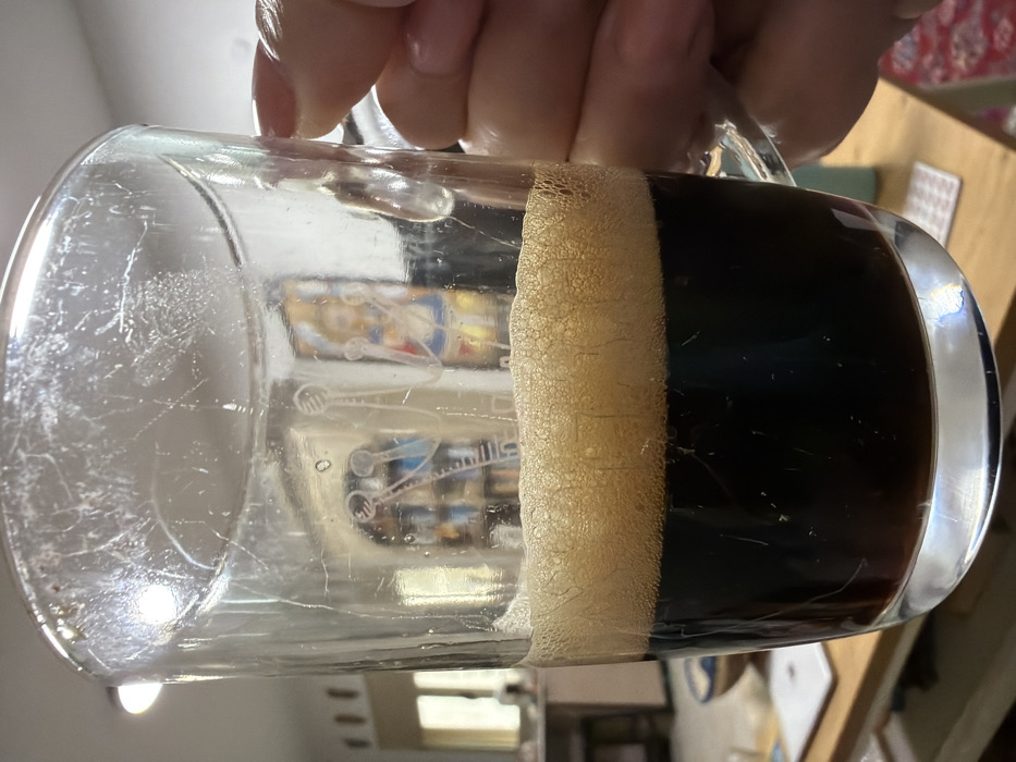 A hand holds a partially consumed dark beer in a glass mug, creating a distinct ring of creamy foam. The mug's surface is etched with visible scratches and an image of space ships. A reflection inside the glass shows a room interior. The beer itself is a deep, opaque black color. A small portion of a wooden table and patterned rug are visible in the background.