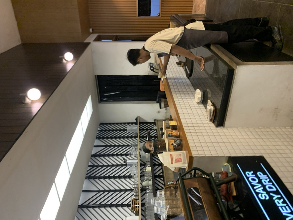 The image shows a cafe interior in Malang, Indonesia, located at -7.939 latitude and 112.631 longitude. A barista wearing a white shirt and black apron stands behind the counter, preparing a drink for a customer who is standing off-camera, holding a cup. The counter is made of black granite and white tiled wall, and behind it are shelves stocked with glass containers of coffee beans and other supplies. The barista is leaning forward, interacting with the customer. The cafe has a modern, minimalist design, with white walls, a black and white geometric patterned wall, and warm wood paneling. The ceiling is white and has recessed lighting. The cafe is clean and well-lit. The focus of the image is on the barista and the interaction with the customer. A sign on the left reads "SAVOR VERY DRIP". The sign on the right reads "FREE WiFi". There are also a few other objects in the image, including a coffee grinder, a shelf with glass containers, and a coffee machine. The cafe seems to be a popular local spot with both the barista and the customer present.