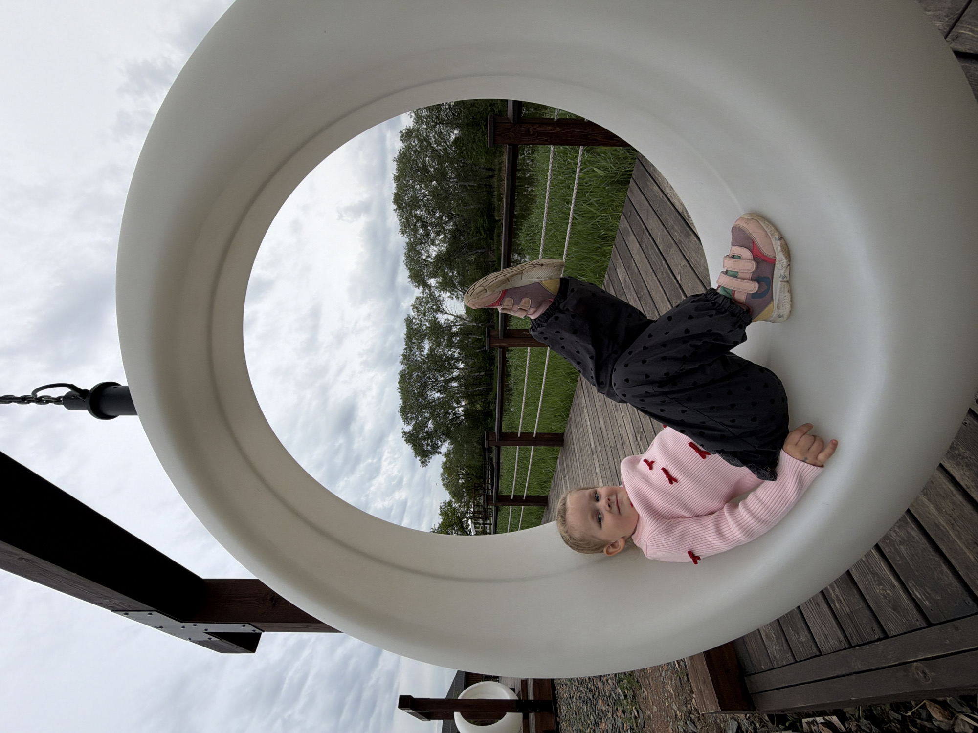 A young child is lying inside a white, circular playground structure in Kamyshin, Russia. The child wears a light pink sweater with small red bow accents, and dark pants with heart-shaped patterns. The child's feet, clad in worn sneakers, extend towards a background featuring a wooden deck, a green grassy area bordered by a low fence, and trees against a cloudy sky. The playground structure is supported by wooden beams. The ground around the structure is covered with wood chips and leaves.