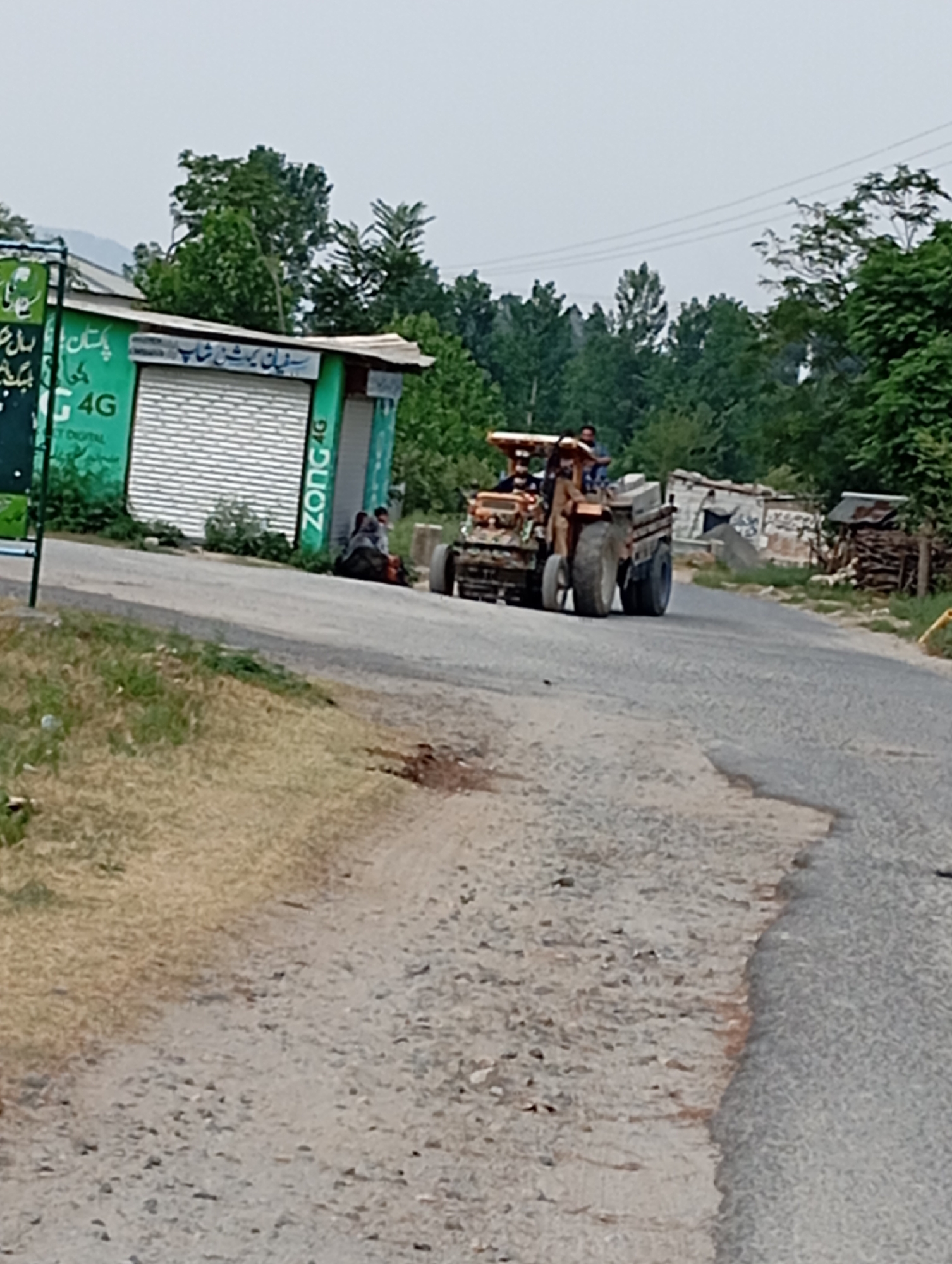 An outdoor, eye-level, medium shot captures a rural scene on a cloudy day. A tractor, pulling a trailer and occupied by three men, travels on a paved road. The tractor is brown and appears to be hauling material in the trailer. To the left of the road is a small, green building with a white roll-up door and signage that includes "4G" and "ZONG". Two individuals are seated on the ground near the building. Behind the building, trees and a partially constructed or dilapidated structure are visible. The foreground consists of a dirt and gravel road.