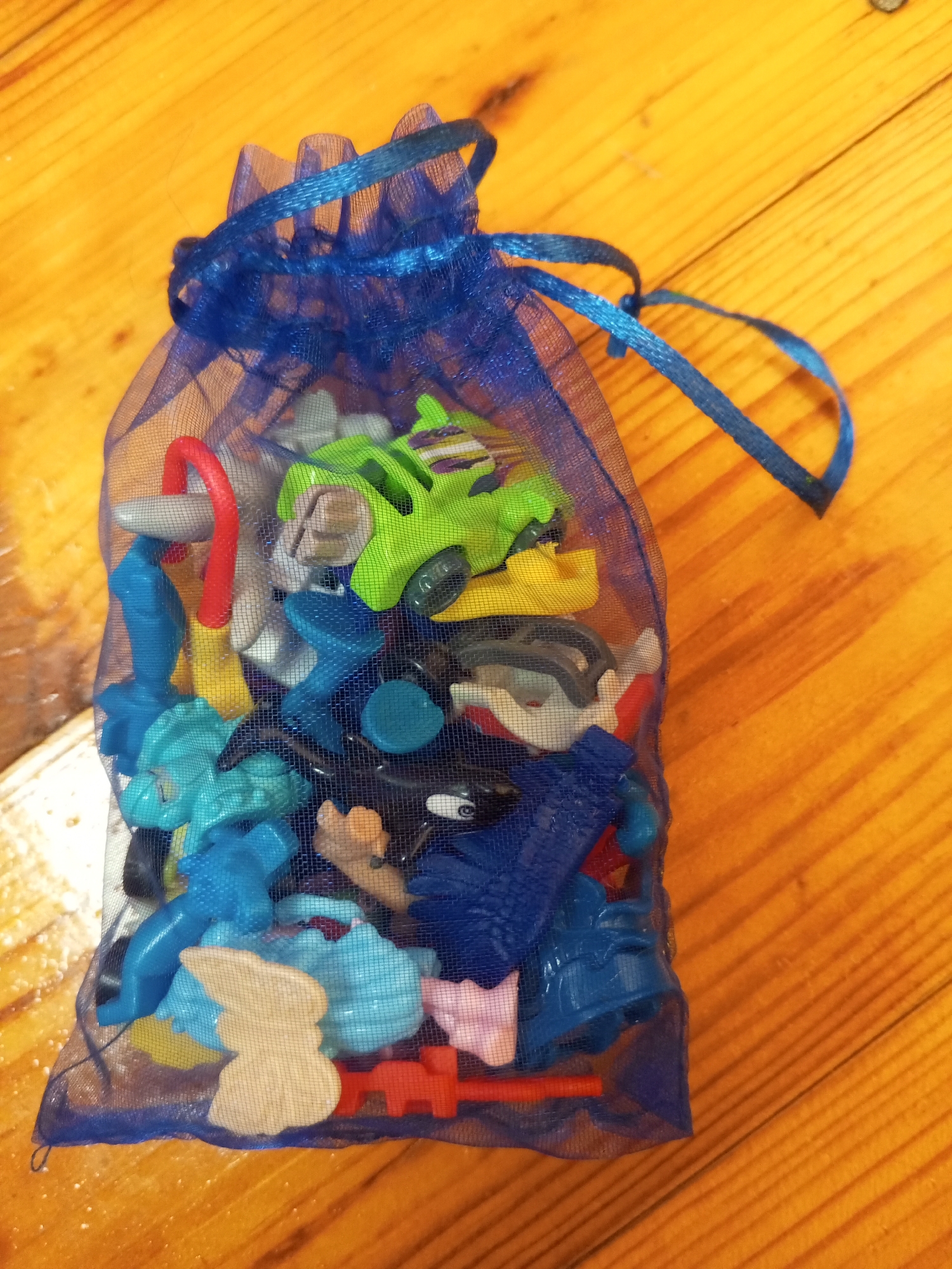 A blue mesh bag filled with small plastic toys sits on a wooden surface. The toys are varied in color and shape, including miniature cars, figures, and other assorted playthings. A blue ribbon is tied around the top of the bag, cinching it closed. The wooden surface appears to be a tabletop or floor, with visible grain and texture. The scene is lit from above, casting slight shadows. No people are present. The image does not contain information about the location in Chistopol, Russia.