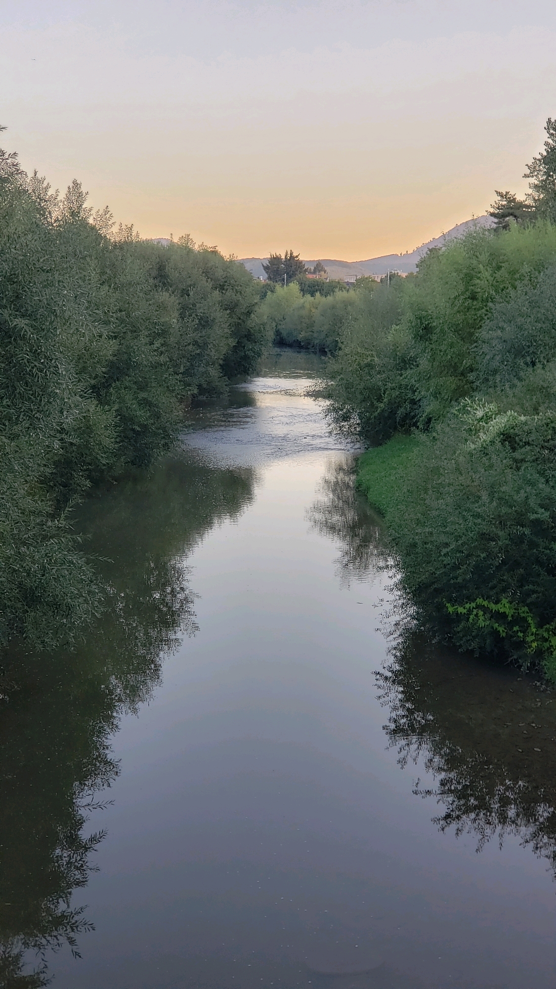 The image depicts a serene river flowing through a lush green landscape, likely at dusk or dawn given the soft, warm light in the sky. The riverbanks are densely lined with trees and bushes, creating a natural corridor. The water's surface is calm, reflecting the sky and surrounding vegetation, with subtle ripples indicating a gentle current. In the distance, rolling hills are visible, suggesting a location with natural beauty. The scene is peaceful and unpopulated. While the image evokes a sense of tranquility, there are no specific landmarks or elements that definitively place it within the city of Prešov, Slovakia without further contextual information.