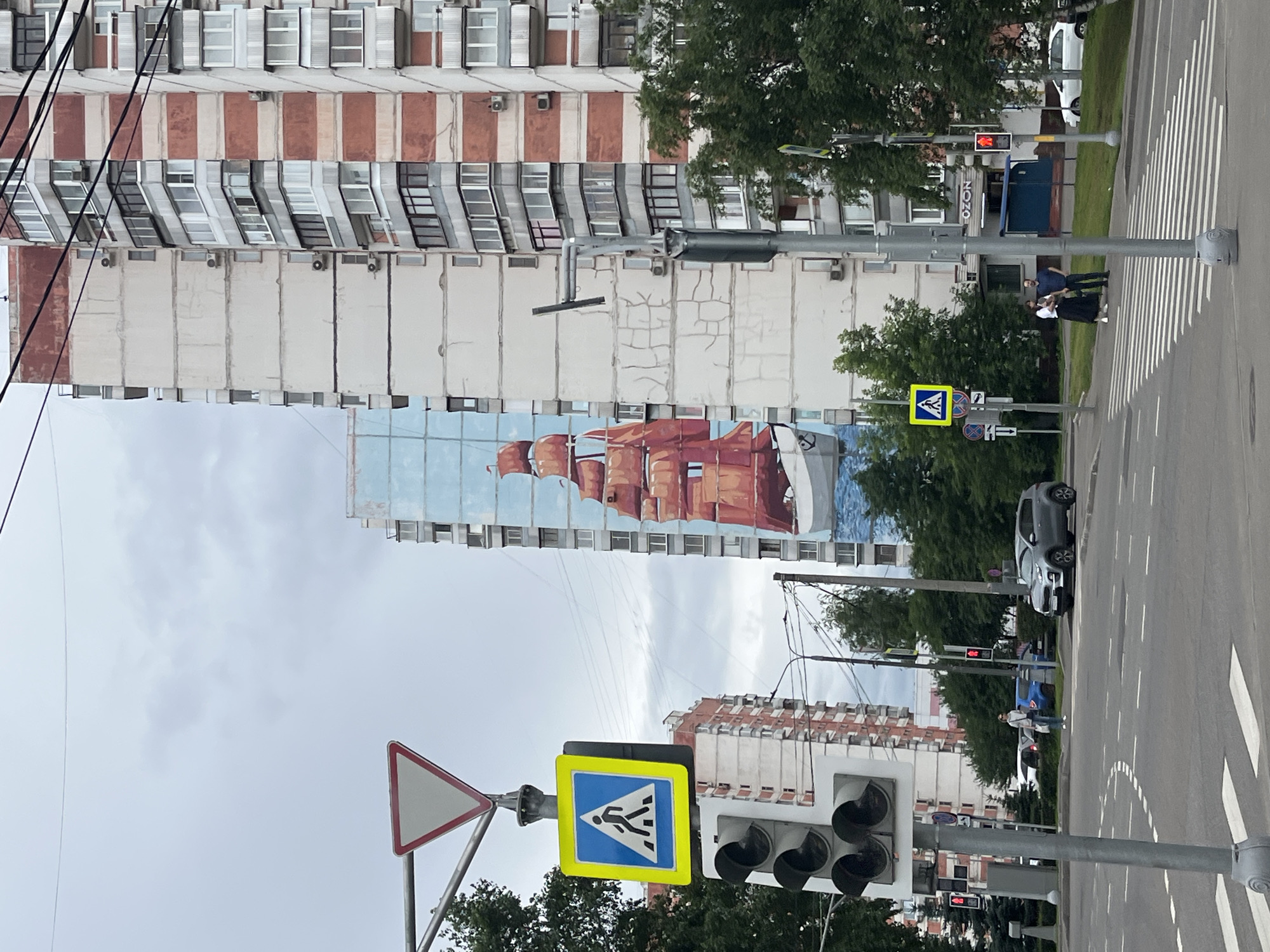 The image depicts a street scene in Moscow, Russia, with a focus on a tall residential building adorned with a large mural of a sailing ship. The mural covers a significant portion of the building's facade, featuring a prominent red-sailed schooner against a light blue sky. In the foreground, a traffic light pole with a pedestrian crossing sign and a yield sign is visible. The street has clear lane markings and a pedestrian crosswalk. Cars are present on the road, including a silver SUV and a blue hatchback. Several people are walking on the sidewalk and near the crosswalk. The background shows other apartment buildings, trees, and power lines under a cloudy sky. The overall environment suggests a typical urban setting in Moscow.
