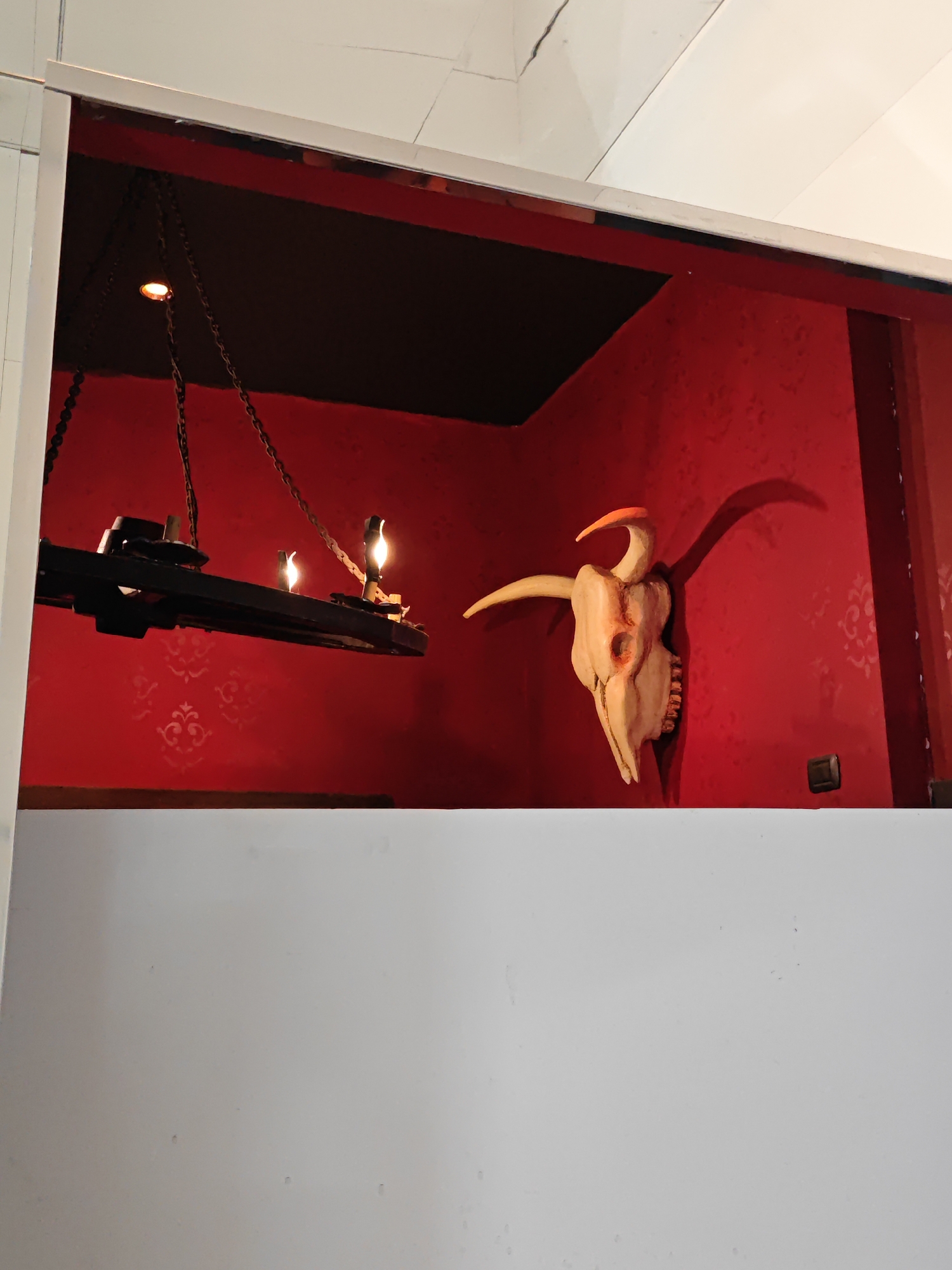 The image depicts an indoor scene with a stark white wall in the foreground and a red wall with a subtle damask pattern in the background. A black, rectangular chandelier with three visible lit bulbs hangs from chains, suspended in front of the red wall. To the right of the chandelier, a light-colored, possibly wooden, animal skull with an orange beak-like protrusion is mounted on the red wall. The skull casts a shadow on the wall. A small dark rectangular object, possibly a light switch or outlet cover, is visible on the lower right side of the red wall. The scene appears to be set in a dimly lit room, possibly a decorative display or an art installation. There is no indication of the city of Turin, Italy, within the image itself.