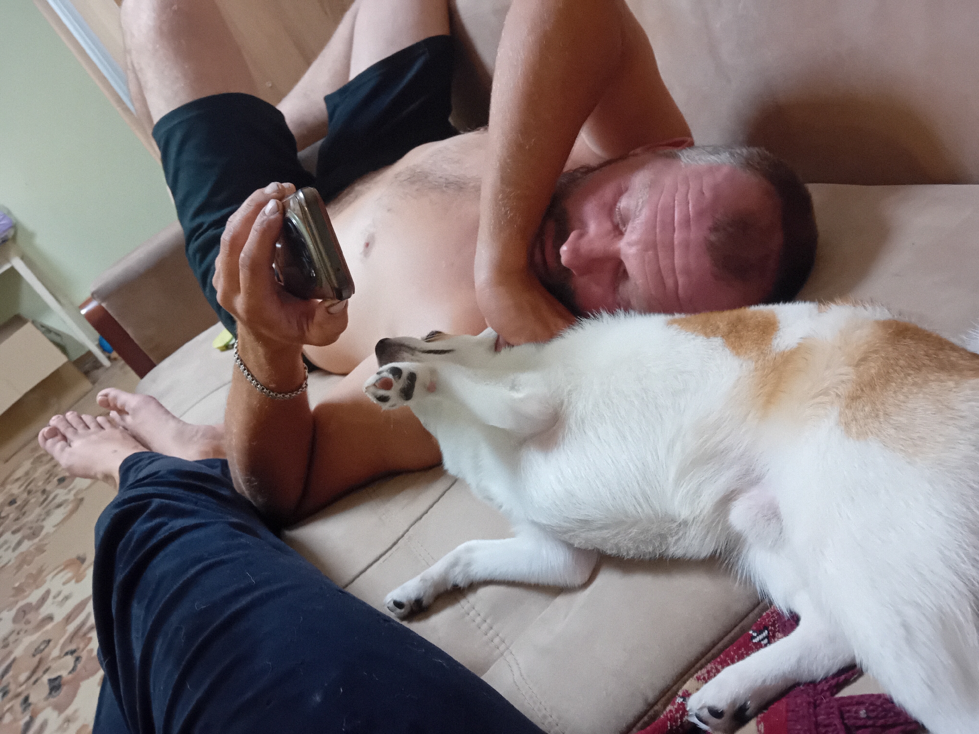 A man is lying on a tan couch, holding a smartphone in his left hand. A small dog, primarily white with brown markings, is lying on the couch next to the man, facing away from the camera. The man is wearing black shorts and the dog appears to be a Shiba Inu or similar breed. The background shows a light-colored wall and a patterned rug on the floor. The scene appears to be indoors, likely in a residential setting.