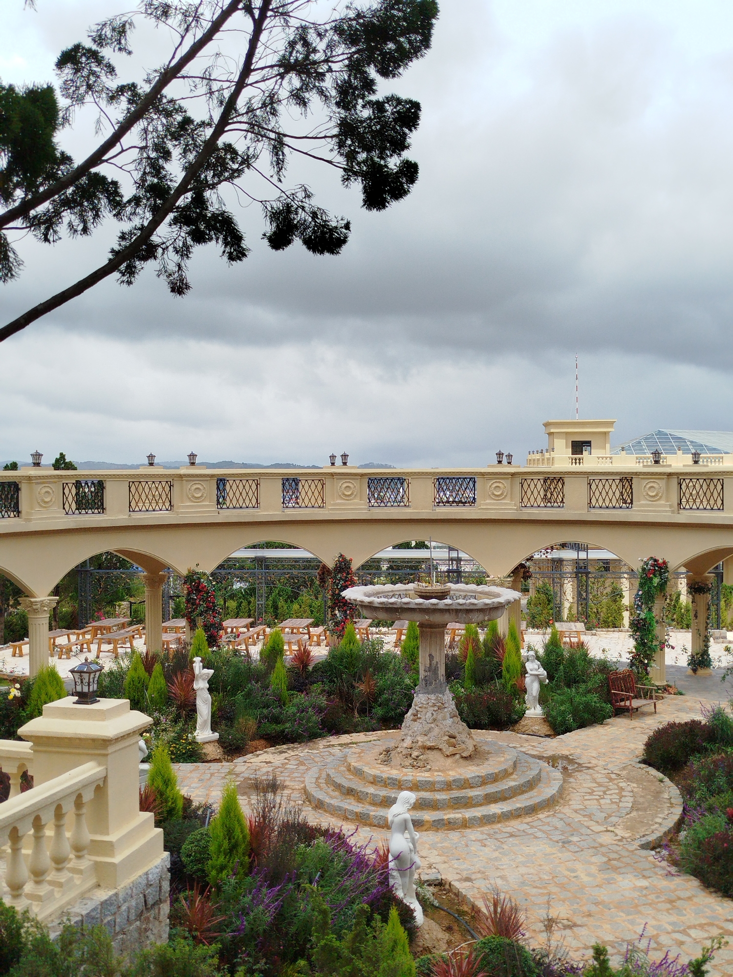 The image depicts a serene garden courtyard in Lạc Dương, Vietnam. The foreground features a decorative fountain surrounded by lush greenery and statues. A paved pathway winds through the garden, leading to an elegant beige archway adorned with railings and lanterns. In the background, several picnic tables with benches are arranged under the archway, suggesting an outdoor dining area. The sky is overcast with dramatic clouds, creating a moody ambiance. The architectural style is reminiscent of European classical design, with columns and ornate details on the archway and surrounding structures.