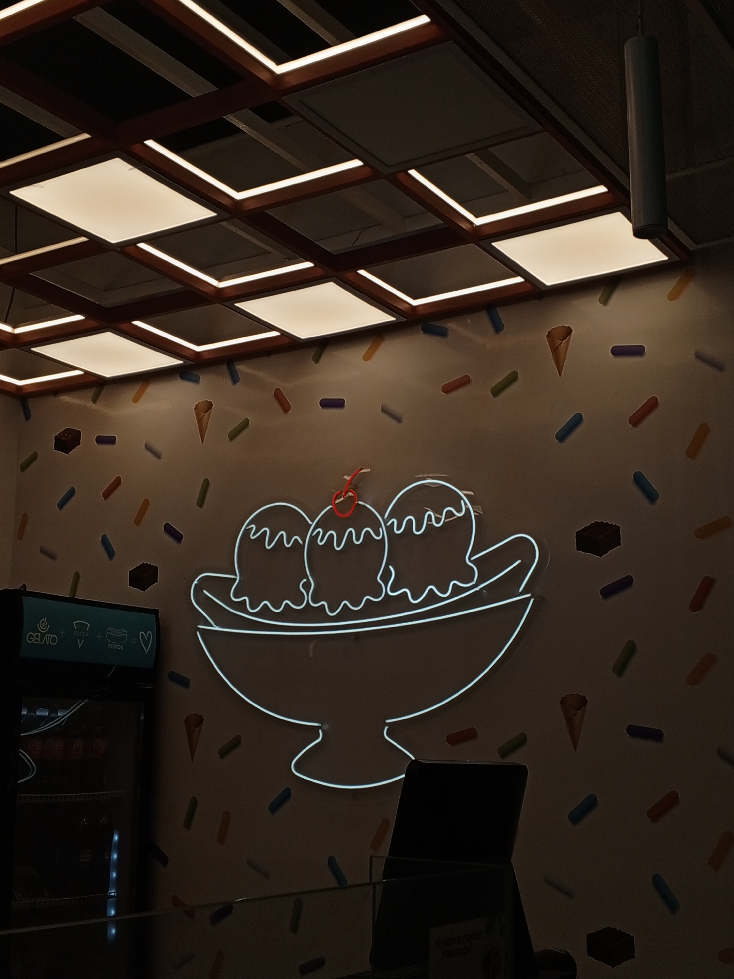 The image presents the interior of what appears to be a dessert shop or gelato parlor, likely at night or during the evening, given the reliance on artificial lighting. The central subject is a vibrant neon sign on a light beige wall, illustrating an ice cream banana split with three scoops of ice cream, wavy toppings, and a red cherry. The wall surrounding the neon sign is whimsically decorated with numerous colorful sprinkles (blue, red, green, purple, orange), cut-outs of ice cream cones, and chocolate bar fragments.
Above, the ceiling features a grid structure with square lights, some illuminated with a warm white glow, while others remain dark, casting a patterned light on the space. The ceiling framework is dark. A dark cylindrical object, possibly a speaker or part of the ventilation system, hangs from the ceiling in the upper right.
On the left side of the frame, a dark display refrigerator or cooler is partially visible. Text on its top panel clearly reads "GELATO" followed by "+ VISTA + PERROS = ❤", suggesting a menu offering or a specific theme. Faint, unreadable text is visible on items within the cooler. The lower right foreground shows a dark, indistinct object, possibly a counter or a screen. The overall ambiance is inviting and playful, designed to attract customers to a sweet treat establishment.