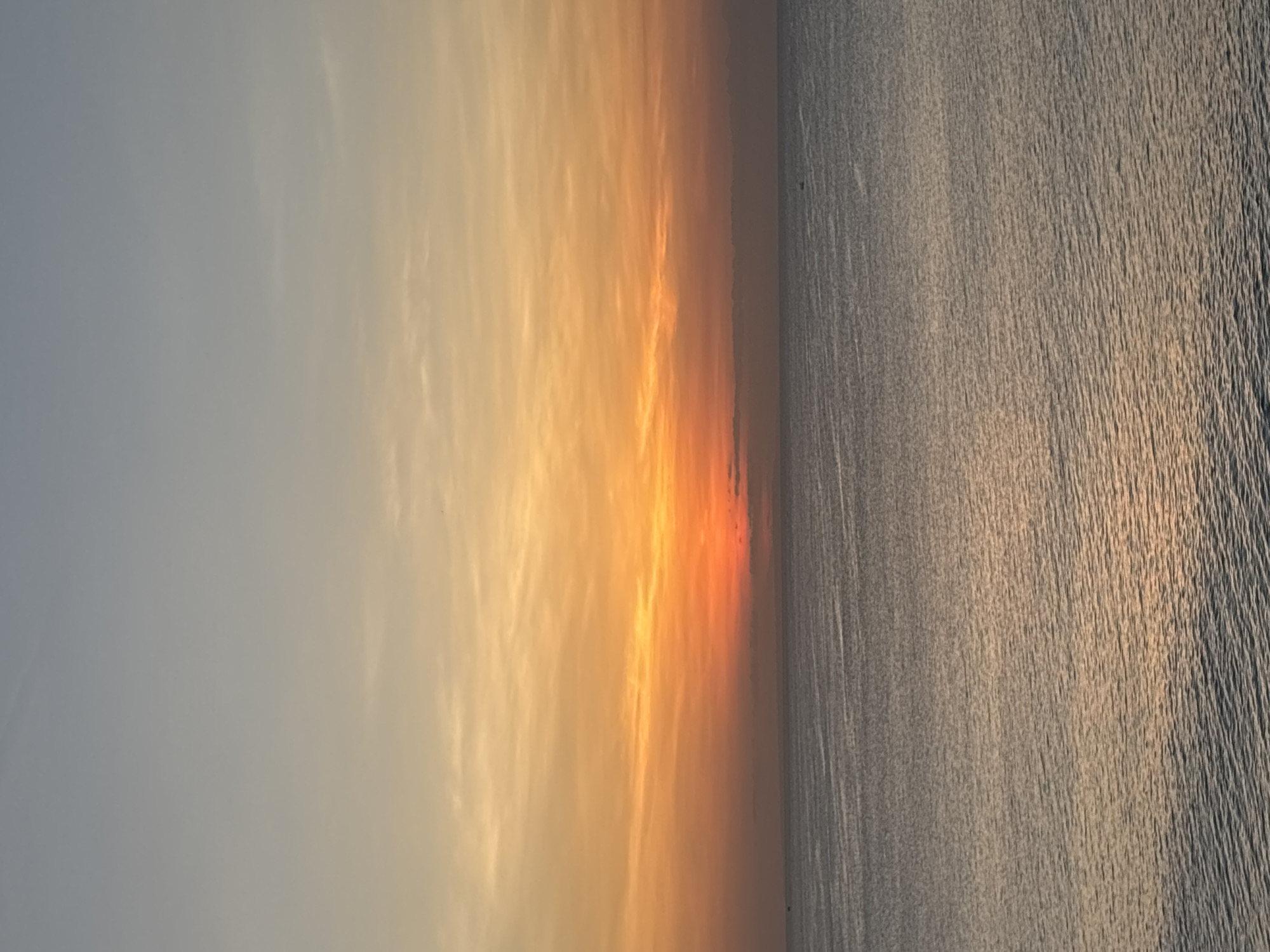 The image depicts a sunrise or sunset over the ocean. The sky transitions from a light grey to orange and yellow. The ocean surface is visible, with ripples and light reflecting. The location is likely Kuta Selatan, Indonesia. No people are visible.