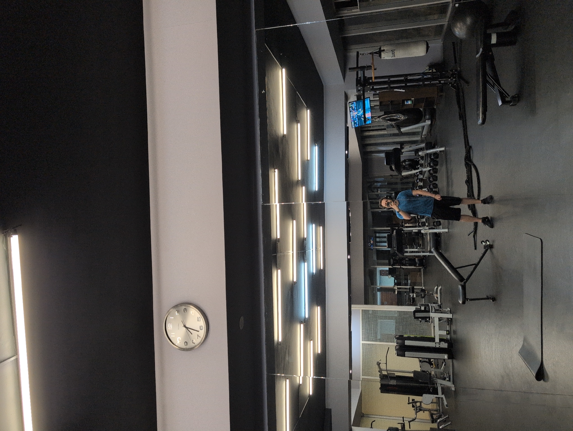 The image depicts the interior of a gym, possibly located in Mexico City, Mexico. A man in a blue shirt and dark shorts is standing in the gym, holding a cell phone. The gym features various exercise machines, including weightlifting equipment and a punching bag. The walls are decorated with linear lights, and a clock on the wall indicates the time. The gym appears well-equipped and organized.