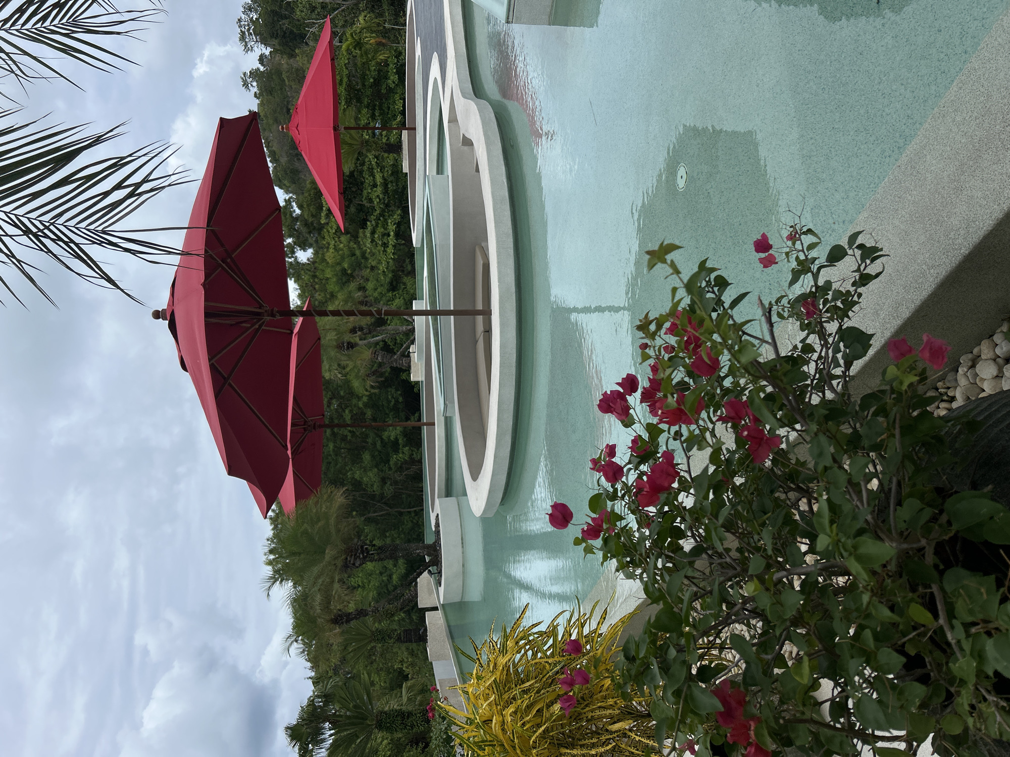 The image captures an outdoor scene in Rawai, Thailand, featuring a multi-tiered swimming pool with light blue water. The pool's edge is adorned with white, curved architectural elements. Red umbrellas are positioned nearby, casting reflections on the water's surface. Lush greenery, including a flowering bush with red blossoms and yellow foliage, frames the pool. Palm fronds are visible in the upper left corner against a backdrop of trees and a cloudy sky. The scene appears serene, suggesting a resort or private residence.