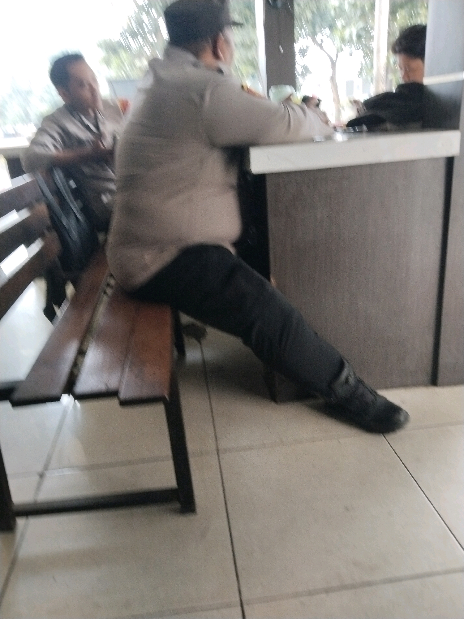 The image shows three individuals in what appears to be an indoor seating area with large windows overlooking an outdoor environment with trees. One individual in the foreground is wearing dark pants and a light-colored long-sleeved shirt and a dark cap. This person is seated on a wooden bench with dark metal legs. The bench is positioned next to a counter with a dark textured surface and a white trim. Another individual, partially visible on the left, is also seated and appears to be wearing a light-colored shirt and dark pants, looking towards the foreground individual. A third individual is visible in the background, seated behind the counter, wearing a dark top. The flooring consists of light-colored square tiles. The overall lighting suggests it might be daytime, with natural light coming from the windows. The context of the image, with the uniforms and seating arrangement, could indicate a waiting area or a casual dining establishment. The specific location in Serang, Indonesia is not identifiable from the visual information alone.