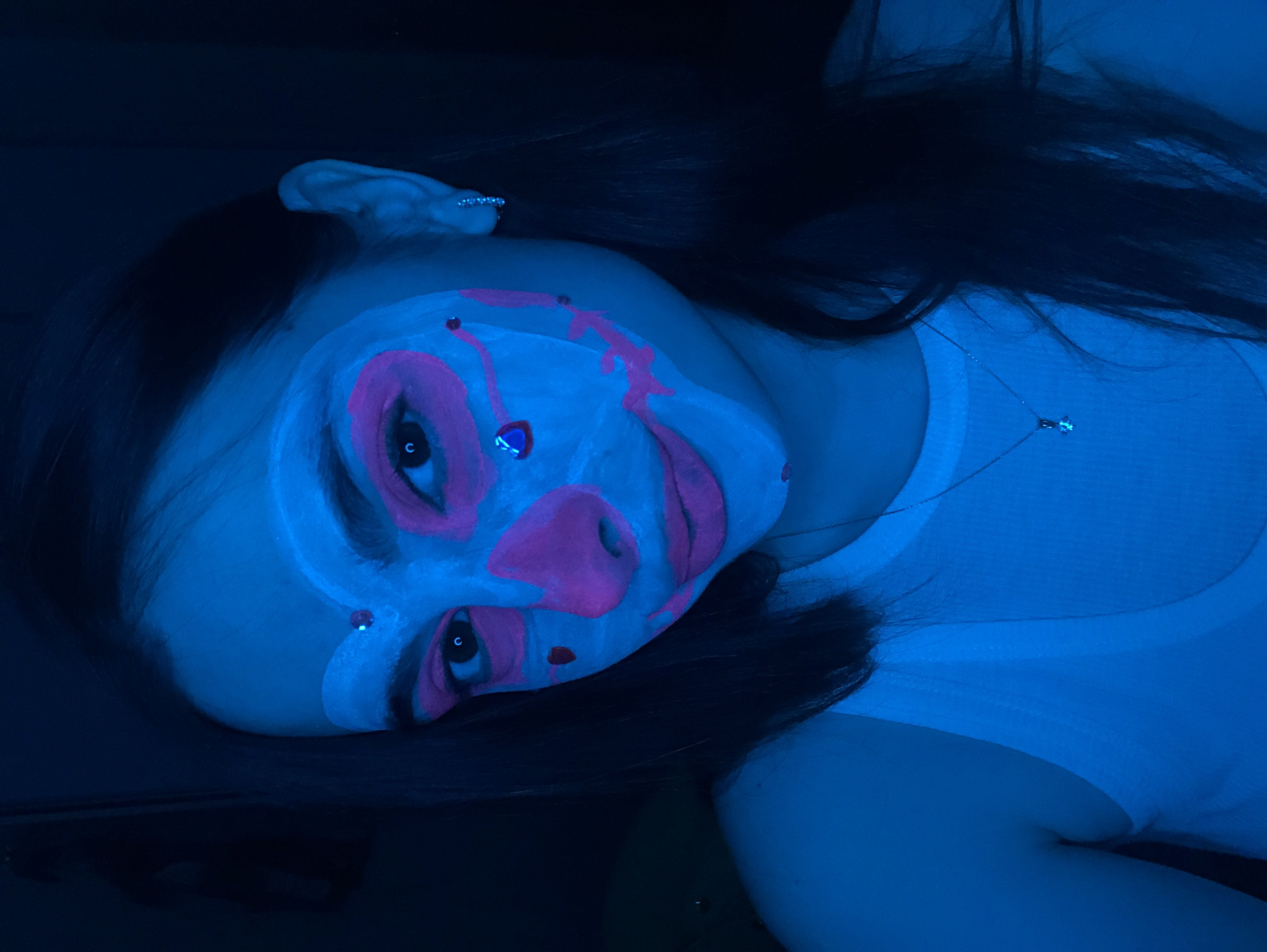 The image captures a close-up portrait of a person, likely female, lying down in a dark setting under strong blue/UV light. The person's face is elaborately painted with a UV-reactive makeup design, predominantly in a glowing light blue or white hue, accented with bright pink details. These pink lines define large, stylized eyes, a clown-like nose, and exaggerated mouth and cheek areas, giving the appearance of a skull or a theatrical character. Abstract pink streaks also run across the forehead and cheeks.
The person's eyes are open, and their pupils are visible. In the reflection of the left eye, a small 'c' shape is discernible, likely an environmental reflection rather than intentional text. Small, sparkling earrings are visible on both ears, and a delicate silver-colored necklace with a small, clear or blue pendant rests on the person's neck and chest, partially glowing under the UV light. The person wears a simple white top or shirt, which also appears to fluoresce under the blue illumination. Long, dark hair is spread out, blending into the dark background.
The overall atmosphere is artistic and somewhat otherworldly, suggesting a performance, a themed party, or a creative photo shoot, possibly related to Halloween or similar events. The intense blue lighting indicates the photo was taken in a dark environment, likely indoors and at night, in Noviy, Russia. The person maintains a neutral to serious expression, enhancing the artistic quality of the portrait.