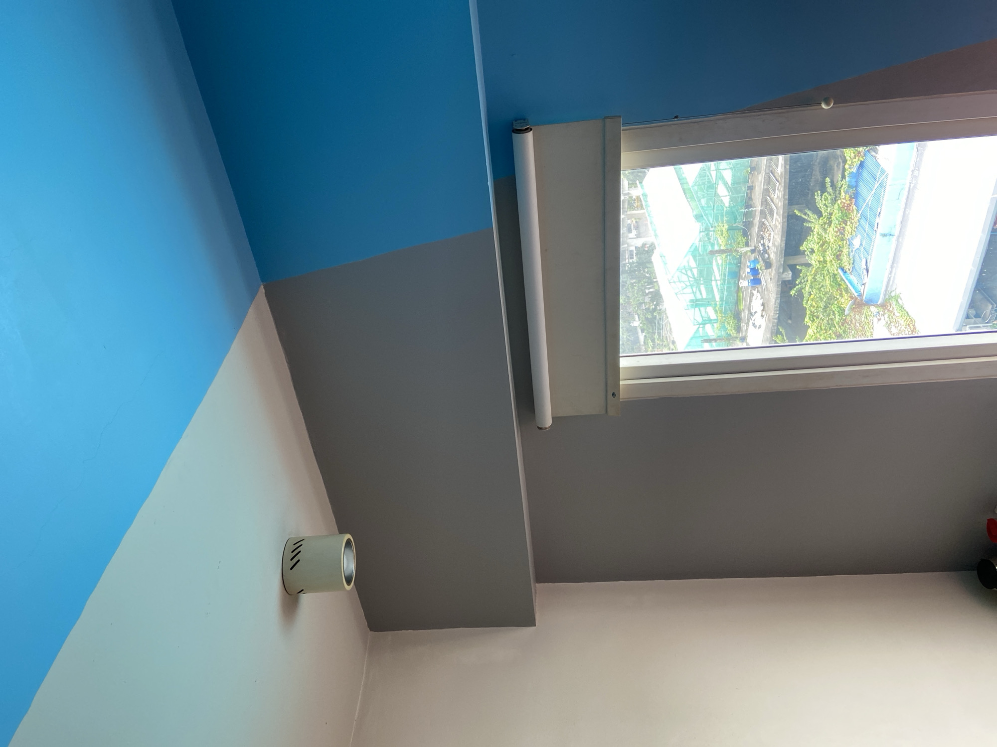 The image shows an interior view of a room with a distinct color-blocked design on the walls and ceiling. The ceiling is split diagonally, with the upper portion painted bright blue and the lower portion a lighter shade. A white cylindrical light fixture is mounted on the lighter section of the ceiling. The walls are also painted in contrasting colors: a light cream color on the left, a medium gray in the center, and a bright blue on the right. A window with a roller blind is visible in the center right of the image. The view outside the window shows buildings, greenery, and what appear to be greenhouses or construction sites, suggesting an urban environment. There are no people visible in the image. The lighting suggests it is daytime. Without further information, the specific location in Central Jakarta, Indonesia cannot be determined from the image alone.