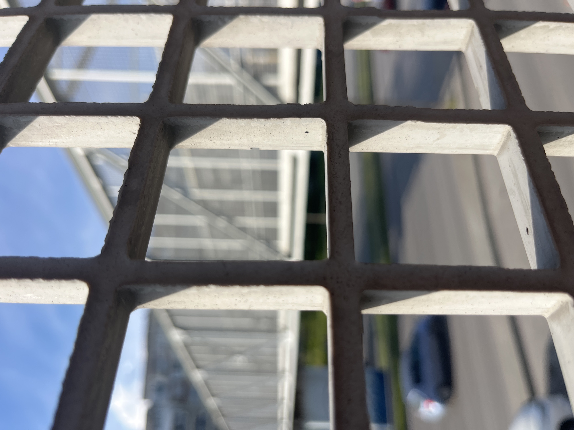 The image shows a close-up view of a metal grid, likely part of a bridge or elevated walkway, with a view of an urban environment visible through the openings. The background includes a structure with white horizontal lines, possibly a staircase or another walkway, a blurred image of a street with a dark car and some greenery. Given the prompt specifies the city of Varna, Bulgaria, the image could potentially be depicting a pedestrian bridge overlooking a street in an area of the city with modern construction.