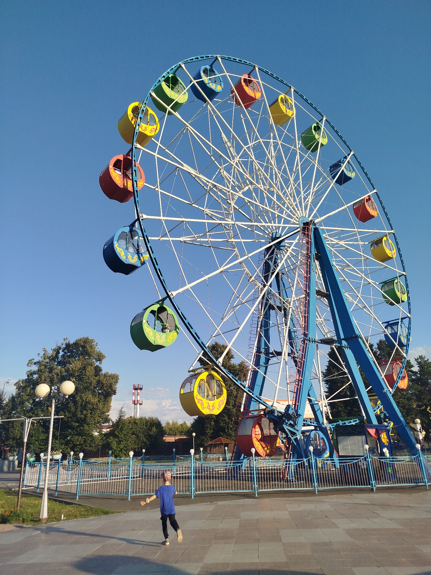 The image depicts a large Ferris wheel in an amusement park located in the city of Yalutorovsk, Russia. The Ferris wheel is colorful, with blue structural supports and white spokes, and its gondolas are painted in various bright colors, including red, yellow, and green, each with decorative yellow accents. The sky is clear and blue, suggesting fair weather. In the foreground, a child wearing a blue shirt and a patterned headscarf is running on a paved area. The child is facing away from the camera, with their arms outstretched. Behind the child and the Ferris wheel, there are trees, fences, and park amenities like lampposts. A tall industrial tower with a red and white structure is visible in the background, behind the trees.