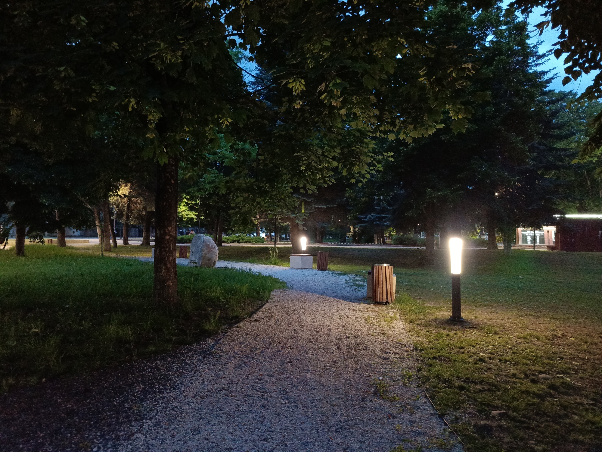 The image depicts a park at dusk in Ust-Dzheguta, Russia. A gravel pathway curves through the scene, flanked by grassy areas and mature trees. Two modern-style lampposts illuminate the pathway and surrounding grass. In the background, a large, light-colored boulder sits on the grass near another lamppost and a wooden trash receptacle. Further in the distance, a building with illuminated windows is visible, suggesting a public facility or establishment. The sky is a deep blue, indicating the transition from day to night. There are no people visible in the image.