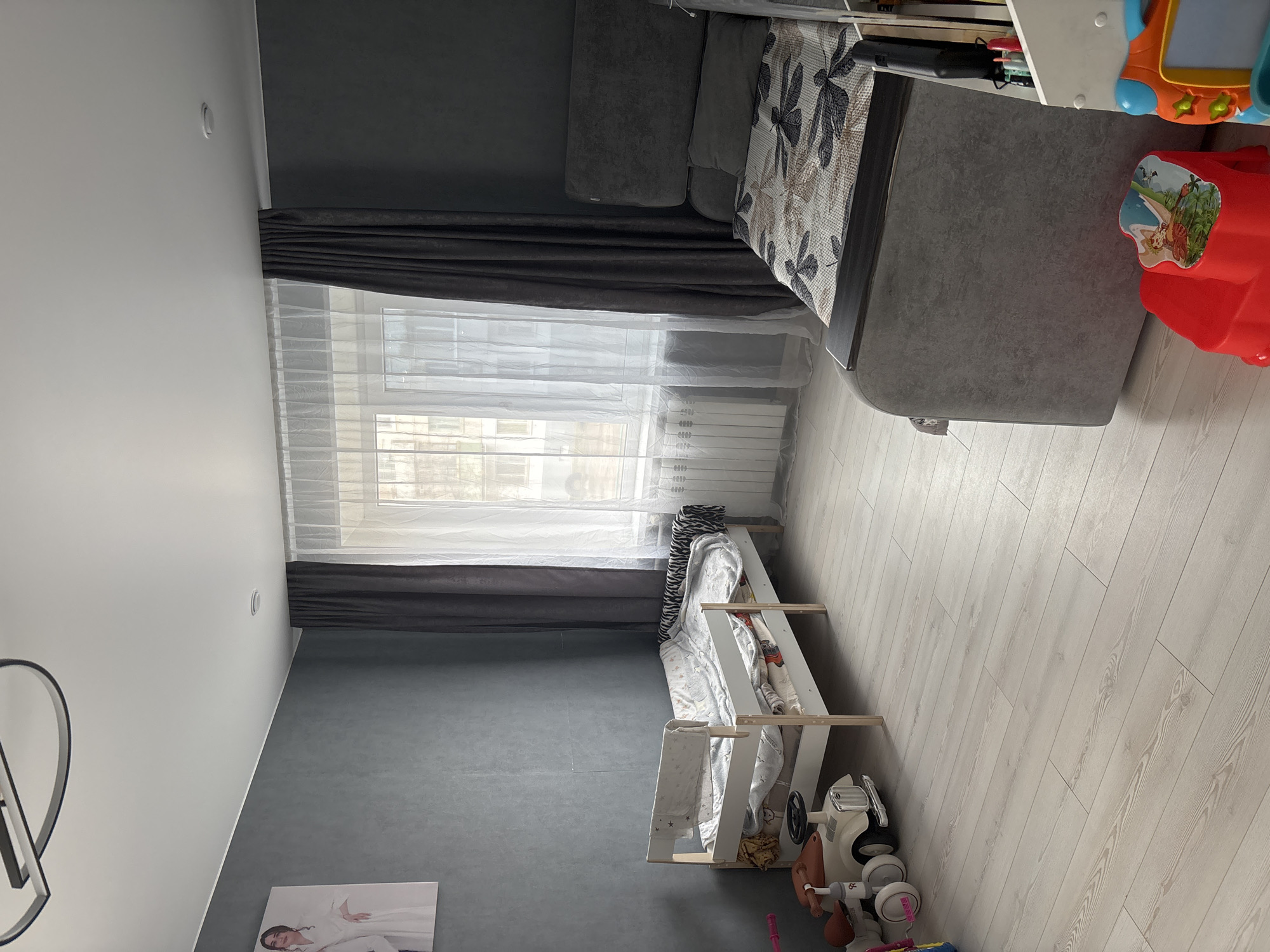 The image shows a room in what appears to be a residential building. A window with white and dark grey curtains is visible, casting light into the room. A grey couch with patterned throw pillows sits near the window. Below the window is a white crib/bed with a patterned blanket. Toys, including a white toy car, are on the floor next to the crib. A framed portrait hangs on the wall, above what appears to be a dark grey wall. The flooring is light wood. The location is Chaikovsky, Russia. There are no people visible.