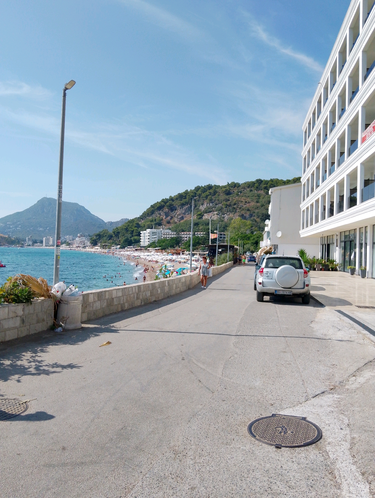 In Sutomore, Montenegro, a silver SUV with a spare tire on the back is parked on the right side of a paved road. The road slopes slightly downward towards the left. A modern, white, multi-story building with arched balconies lines the right side of the road. On the left side of the road, a stone wall separates the paved area from a beach with many people sunbathing and swimming in the blue sea. A tall street lamp stands near the stone wall. Behind the beach, a hilly landscape is covered with lush green vegetation and dotted with buildings. The sky is clear blue with a few wispy clouds.