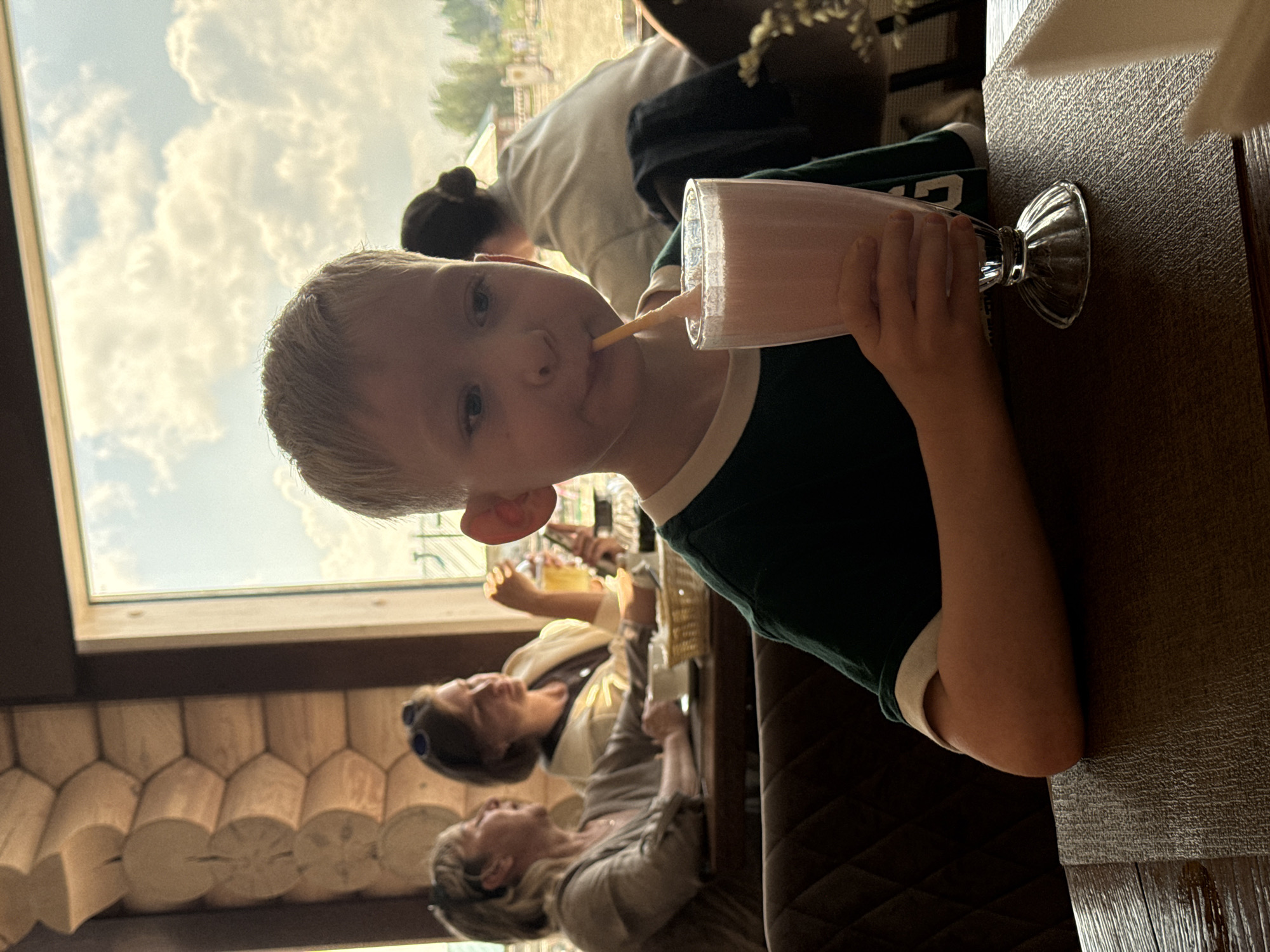 A young Caucasian boy with blond hair is seated at a table in a restaurant, holding a glass of pink milkshake and drinking it with a straw. He is wearing a dark green t-shirt with white trim. In the background, two women are seated at the same table, and another person with dark hair is visible further back. The restaurant interior features a wooden wall and a large window looking out onto a cloudy sky and a landscape that appears to be a farm. The lighting suggests it is daytime. The overall setting is casual and appears to be a family outing.