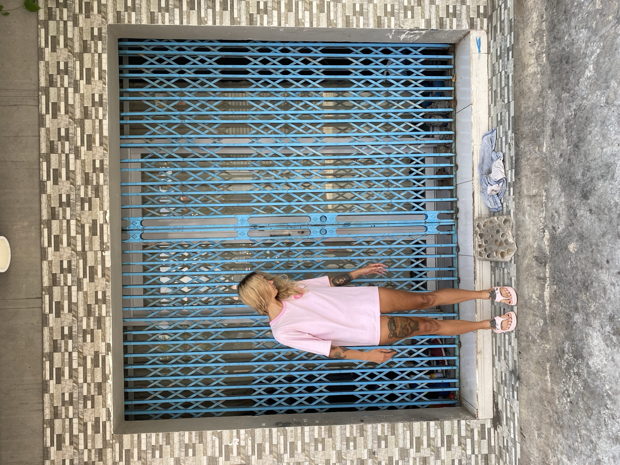 A photograph depicts a blonde-haired woman lying on a closed, blue, metal roll-up security gate in front of a building. The woman is wearing a pink t-shirt and pink sandals. She has tattoos on her arms and legs. The building facade is decorated with a geometric pattern of rectangular tiles. The background suggests an urban environment, possibly in Đồng Nai, Vietnam. A concrete sidewalk runs alongside the building, and a piece of crumpled cloth and a concrete block with holes are present. The photo is taken from an overhead perspective.