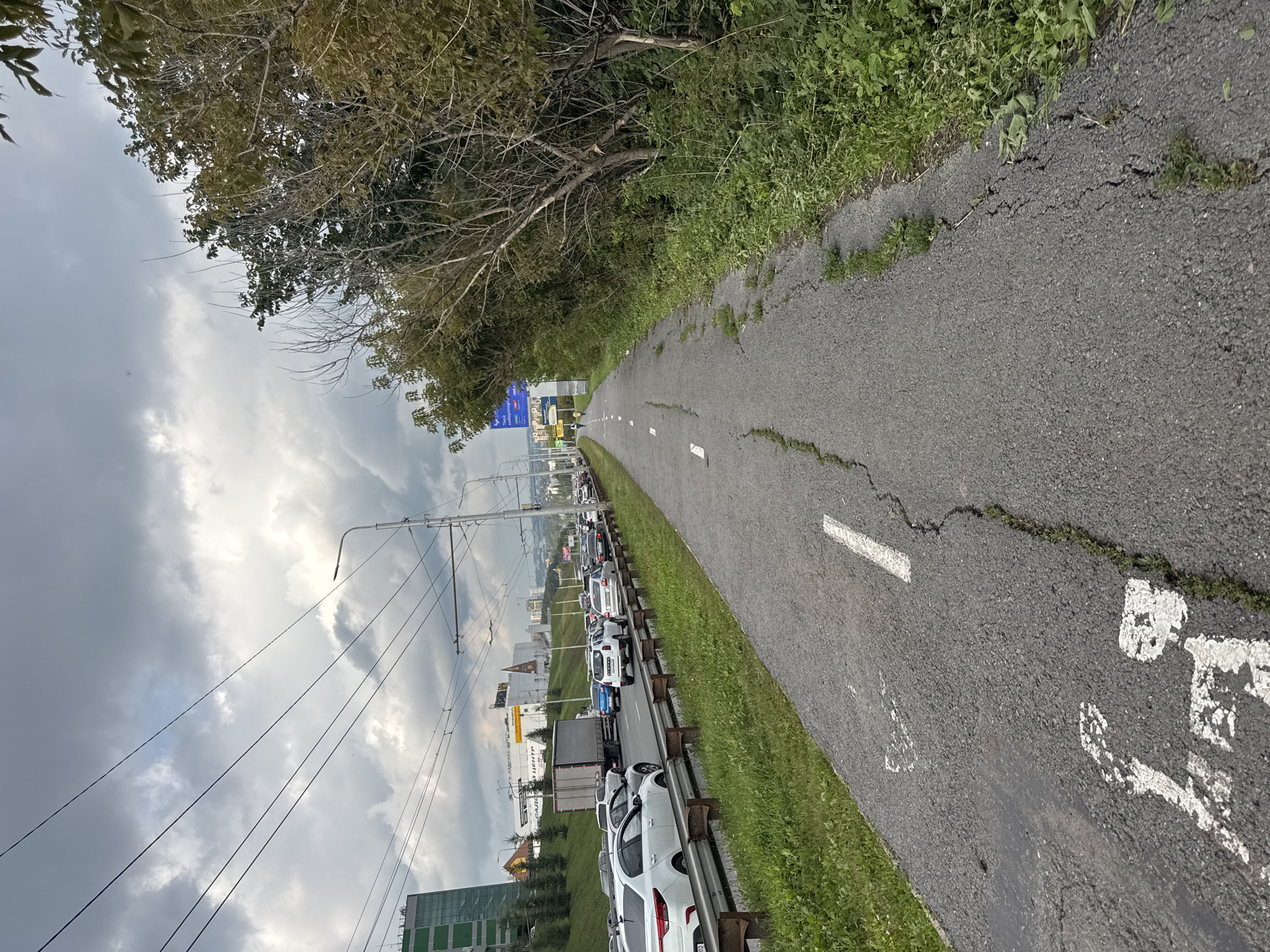 The image depicts a paved bike path with white markings, cracked asphalt, and overgrown grass on the right side. A traffic jam of cars is visible on the left, separated from the bike path by a metal barrier. In the background, there are buildings, trees, power lines, and streetlights. The sky is overcast with dark clouds. The scene appears to be captured in a suburban or industrial area, possibly near a highway, based on the presence of traffic and large commercial buildings. The overall impression is one of urban infrastructure juxtaposed with elements of nature. The image appears to be taken in Ufa, Russia, given the specific building architecture and signage visible in the background.