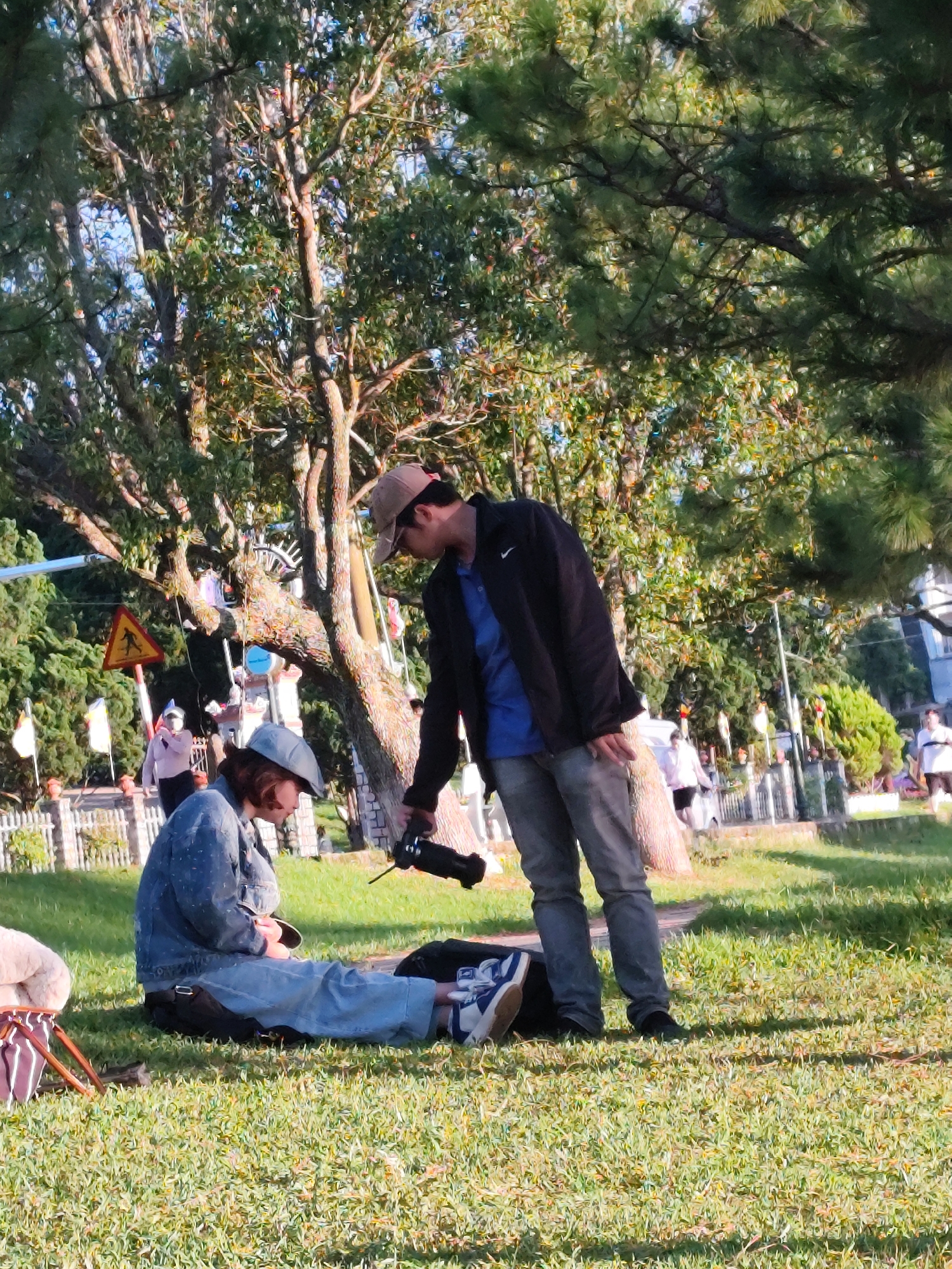 In Da Lat, Vietnam, a man wearing a dark jacket over a blue shirt and grey pants, along with a baseball cap, is holding a camera and appears to be taking a photo of a person sitting on the grass. The seated person, wearing a denim jacket, blue jeans, a blue cap, and white sneakers, is looking down at something in their hands. They are positioned on a grassy area under the shade of large trees. In the background, a white picket fence encloses a pathway with several people walking and standing. A yellow triangular road sign depicting a pedestrian crossing is visible to the left. The scene suggests an outdoor recreational setting, possibly a park or public garden in Da Lat, given the characteristic lush greenery and trees.